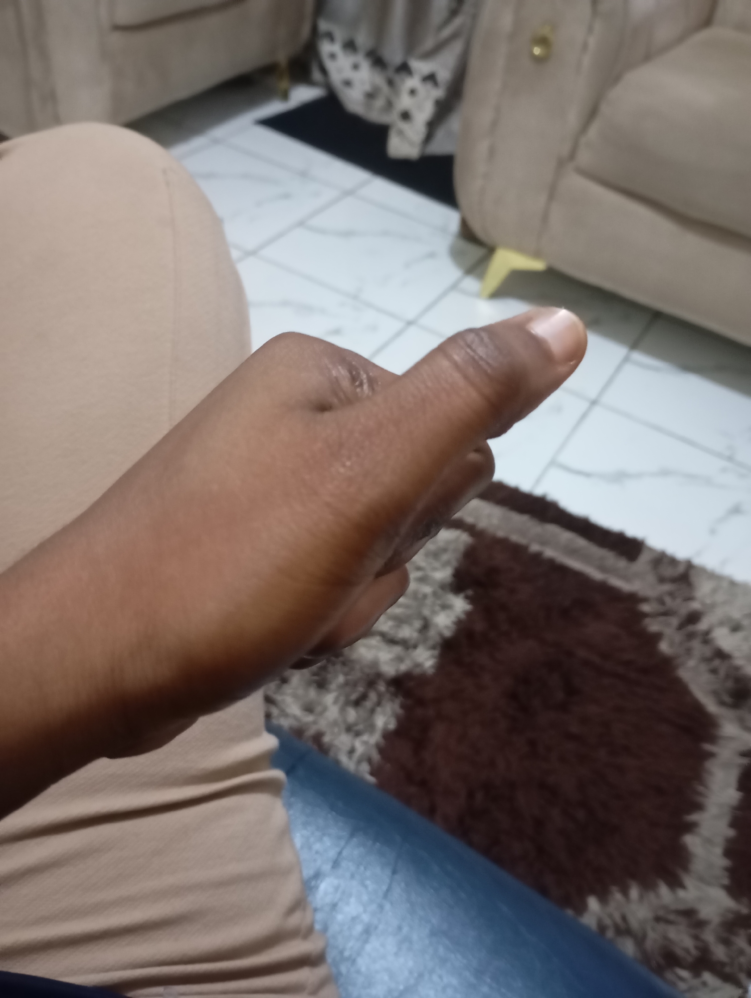 An unidentified individual's hand with dark skin is shown in the foreground, with the thumb extended upward and slightly bent. The hand is positioned in front of a light-colored, ribbed fabric, possibly clothing. In the background, a tiled floor with a pattern of light and dark squares is visible, along with a portion of a cream-colored sofa and a dark brown rug with lighter gray accents. The scene appears to be indoors, in what could be a living room.