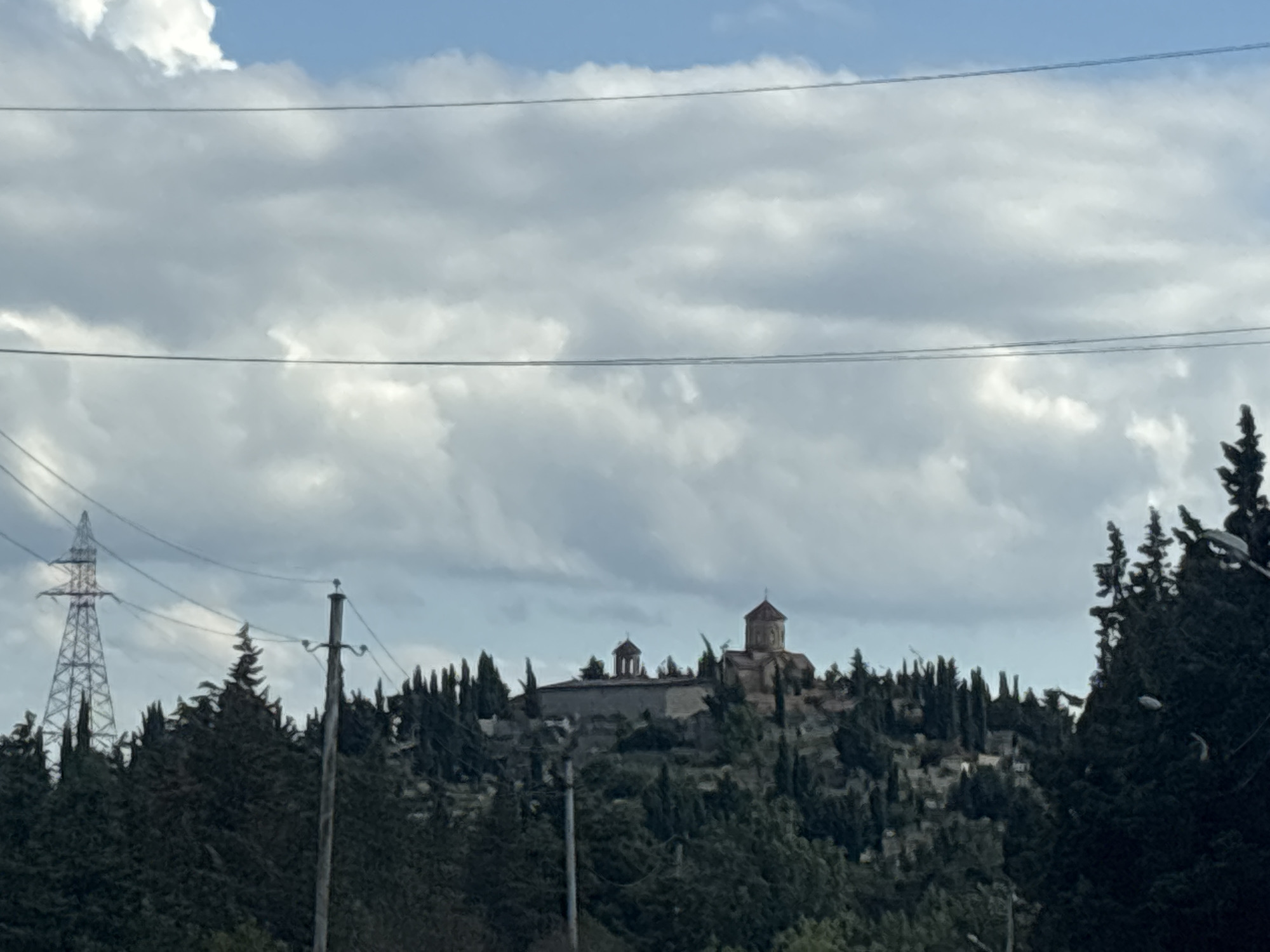 The image shows a landscape view of a hillside covered in trees, with a building situated on the summit. The building appears to be a religious complex, possibly a monastery or church, with multiple structures and domes. The hillside is densely populated with evergreen trees, creating a dark green texture. In the foreground, there are power lines stretching across the frame, with a tall, lattice-like electrical tower visible on the left side of the image. A utility pole with wires is also present in the mid-ground. The sky above is partly cloudy, with patches of blue sky visible between the white and grey clouds. The lighting suggests it is daytime, possibly late afternoon, given the diffused light and cloud cover. The scene evokes a sense of historical or spiritual significance due to the presence of the religious building on the hilltop, contrasted with the modern infrastructure of power lines. The overall mood is tranquil yet somewhat imposing, with the dark foliage and the towering religious structures.