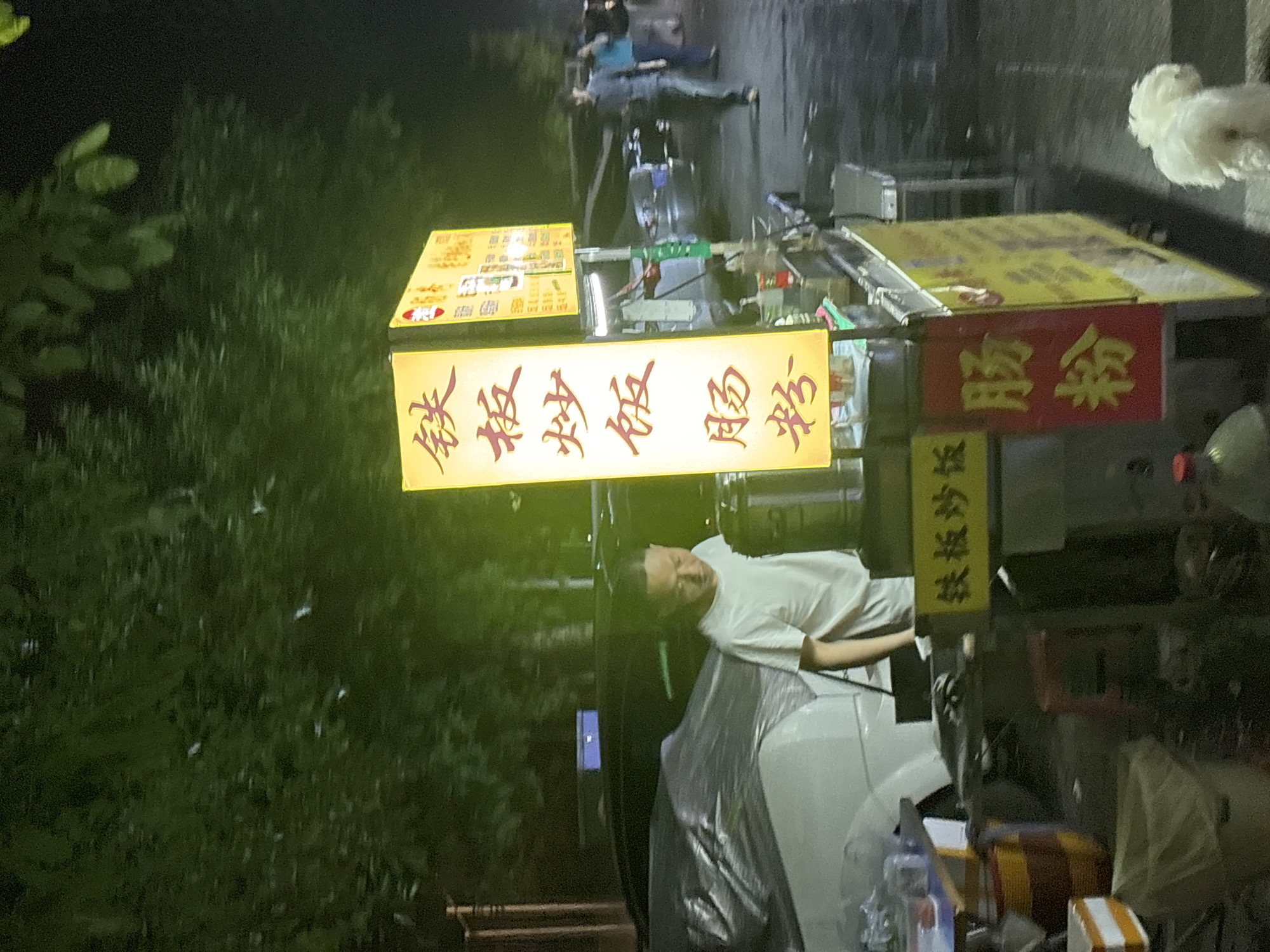 Here's a detailed description of the image:
**Location:** The image appears to be taken at night in an outdoor setting. Given the text visible on signs, the presence of a person, and the style of the objects, it is likely in Nan'an District, China.
**Environment:** The scene is illuminated by artificial light sources, casting shadows. Trees and vegetation are visible on the left side. The ground appears to be a paved surface, possibly a sidewalk or street.
**People:** One person is visible, a man wearing a white t-shirt. He appears to be near the food cart, possibly the vendor. Other individuals are barely visible in the background.
**Objects:** The most prominent objects are food carts. There are multiple carts with signs, possibly indicating the food being sold. The signs feature Chinese characters. Other objects near the food carts include what appear to be cooking equipment, food containers, and potentially a car covered with a cloth.
**Actions and Interactions:** The man in the white t-shirt is standing near the food cart, possibly engaging in work. Other people are near the food cart.
**Overall Impression:** The image captures a nighttime street food scene. The presence of the food carts, the lighting, and the surrounding environment create an impression of a bustling, informal atmosphere.