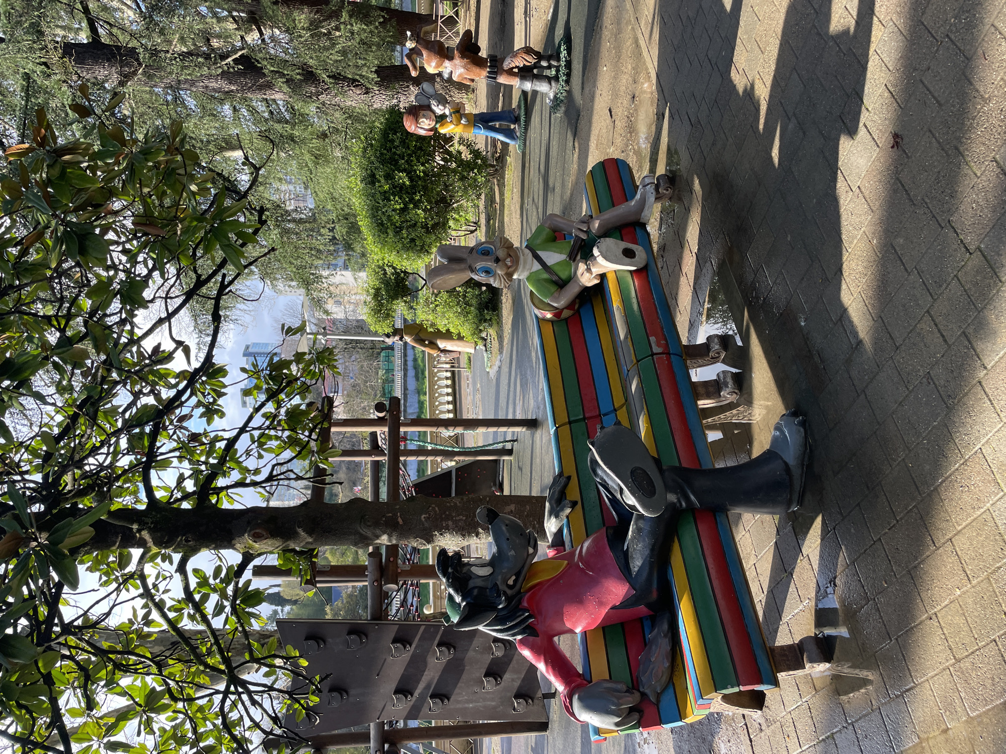 The image depicts a public park in Sochi, Russia, featuring a collection of cartoon-themed sculptures. The sculptures are placed along a paved walkway and on a colorful park bench.
Notable sculptures include cartoon characters. The character on the bench is in a reclining position with a red shirt and black pants, while other sculptures are standing behind the bench. The environment includes trees, shrubs, and other park elements such as a climbing structure visible near the sculptures. The ground surface is wet, suggesting it has recently rained or been watered. There are shadows cast on the pavement, which are likely from the figures. The photograph is taken during daylight.
