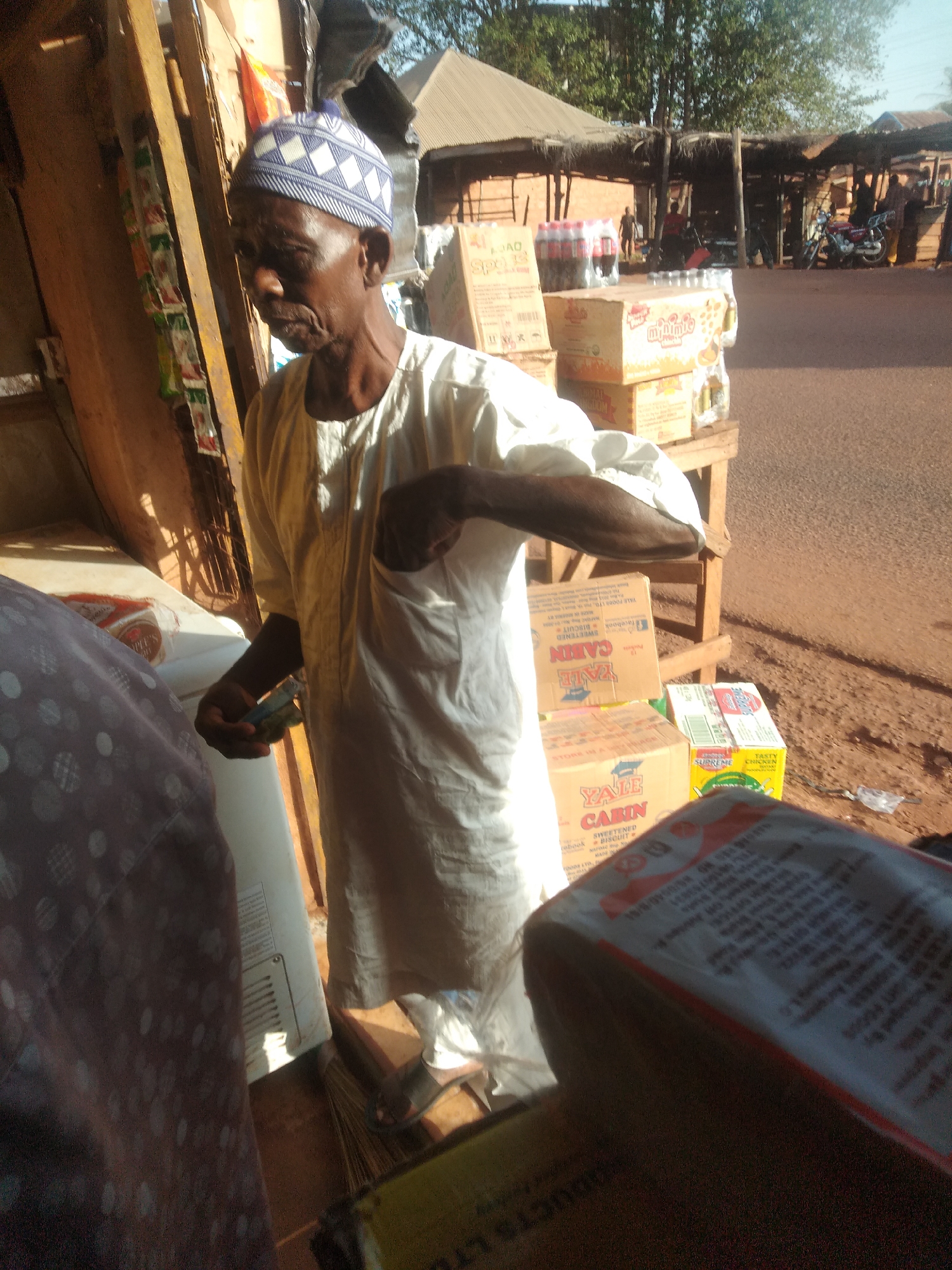 The image appears to be taken in Ejule, Nigeria, showing an elderly man wearing a white tunic and a patterned blue and white cap. He stands next to a cooler, with money in his hand. Behind him, shelves are stocked with various products, including biscuits, packaged drinks, and other goods. A dirt road stretches in the background, with people, motorcycles, and buildings visible. The overall scene suggests a street-side shop or market.