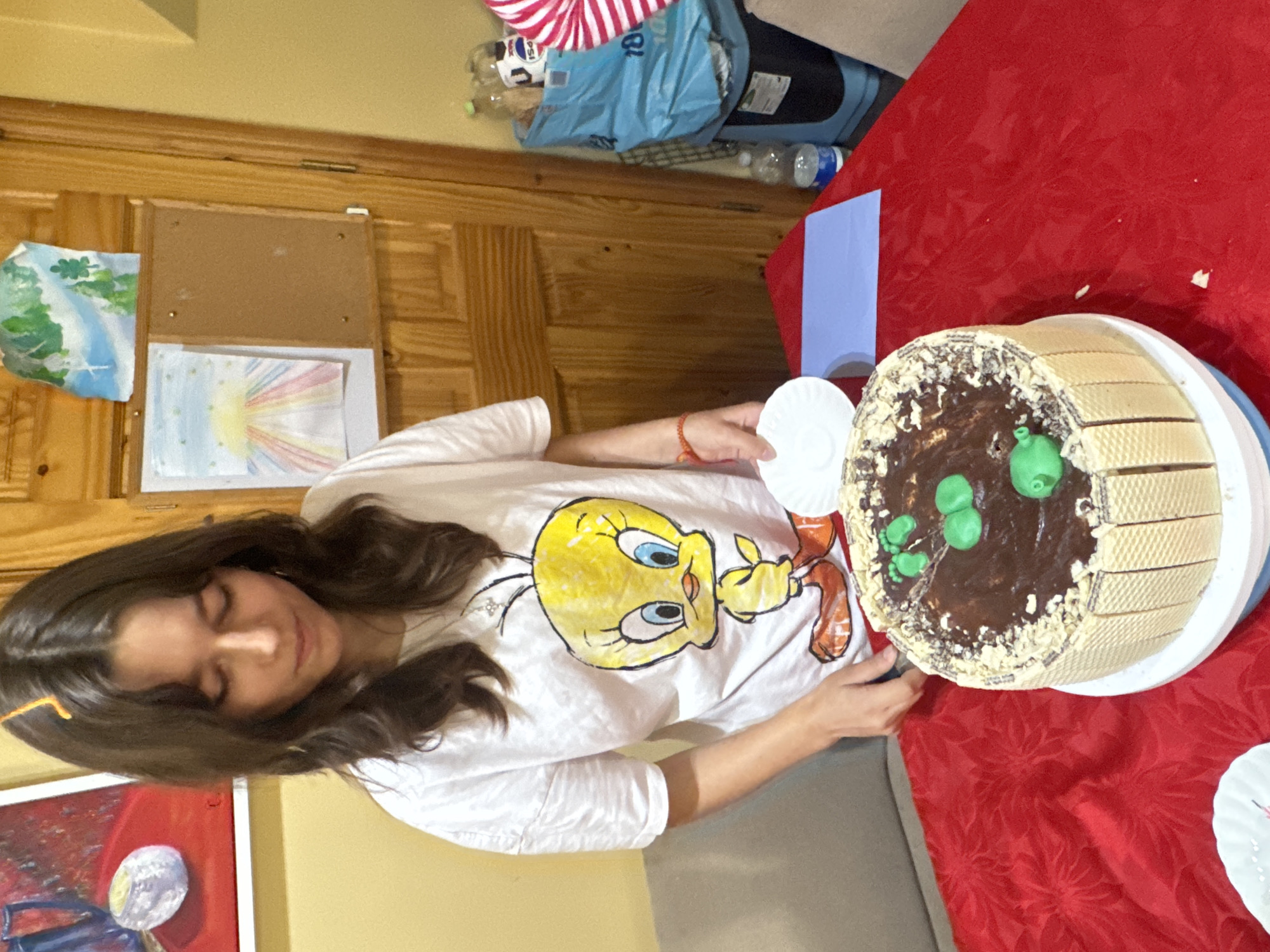 A young woman wearing a white t-shirt with a Tweety Bird graphic stands behind a chocolate cake. The cake is decorated with green fondant, resembling feet and balloons. The cake is surrounded by wafer cookies. The woman is holding a small white plate and is in the process of serving the cake. The scene appears to be indoors, possibly a home setting, with a red tablecloth covering the table. In the background, there are wooden doors and some artwork on the wall, including a painting of a landscape and another drawing. There are also some plastic bottles and a blue bag visible in the background. The lighting suggests it is daytime. There is no visible indication of the location being in Waterford, Ireland.