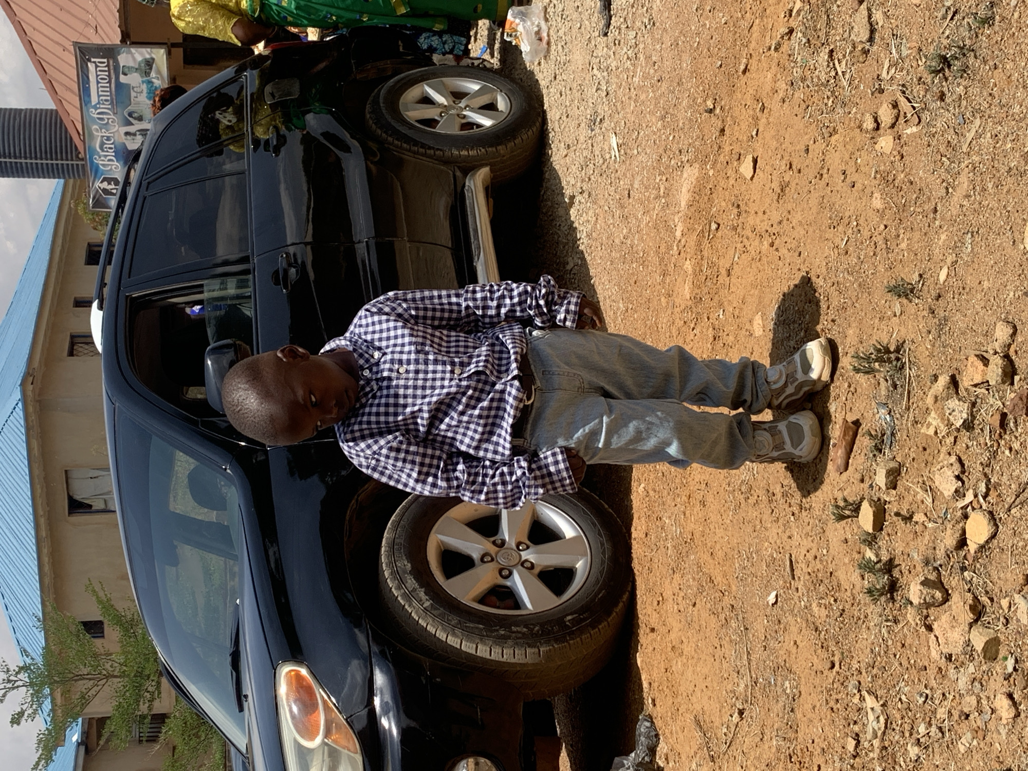 A young boy in a blue and white checkered shirt and jeans stands next to a black SUV, possibly a Toyota Land Cruiser Prado, in Gura Top, Nigeria. The vehicle is partially parked on a dirt surface with small rocks and vegetation. A building with a blue roof and a sign that says "Black Diamond" are visible in the background. The setting appears to be a sunny, outdoor environment.