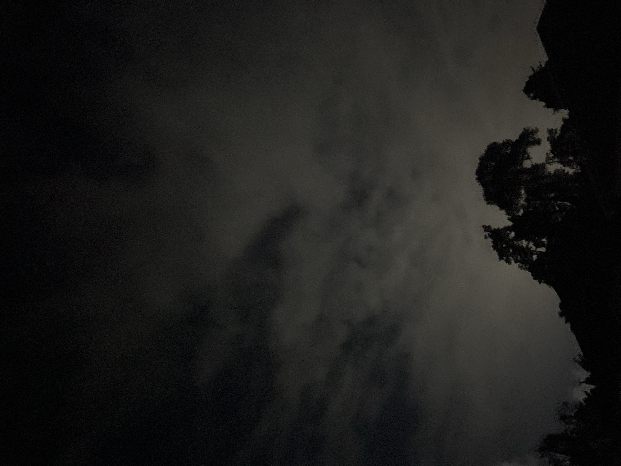 A low-angle shot of the sky at night. Dark clouds fill most of the frame. Silhouetted trees and the edge of a building are visible on the right. The location is unspecified.