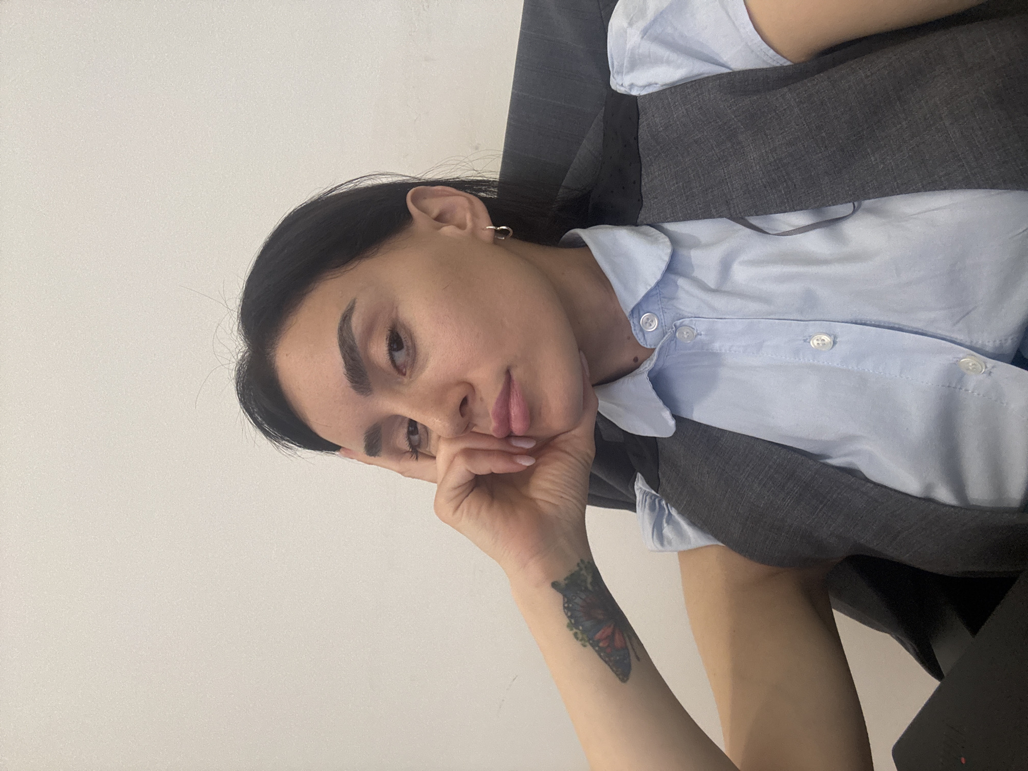 A woman with dark hair is seated indoors. She is wearing a light blue collared shirt with white buttons and a dark gray vest. Her face is resting on her hand, which has a butterfly tattoo on the wrist. The background is a plain white wall. There is no indication of the location being Tbilisi, Georgia.