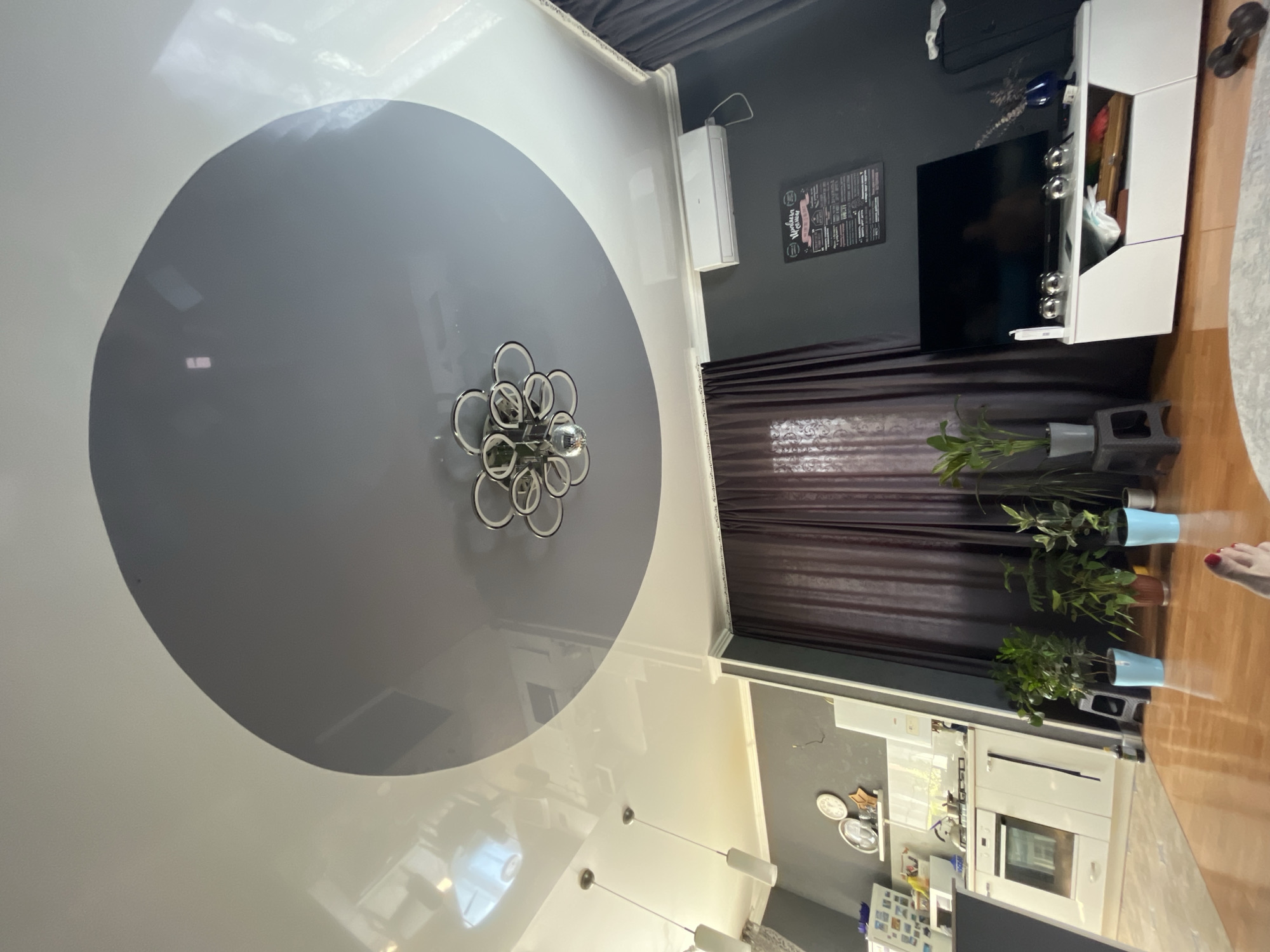 This image depicts an indoor scene, likely a living room and kitchen area, with a view towards a modern ceiling. The ceiling features a two-tiered design: a glossy white outer layer and a matte gray inner circle. A contemporary chandelier with multiple silver rings and a central mirrored sphere hangs from the gray section. On the wall, an air conditioning unit is mounted above dark brown curtains. Below the air conditioner, a dark gray wall displays a poster with text and illustrations, and a flat-screen television is set on a white media console. Several potted plants are placed on the floor and a small stool. To the left, a kitchen area is visible with white cabinets, an oven, and a countertop. The flooring appears to be polished wood. No specific details pinpoint the location to Saratov, Russia.