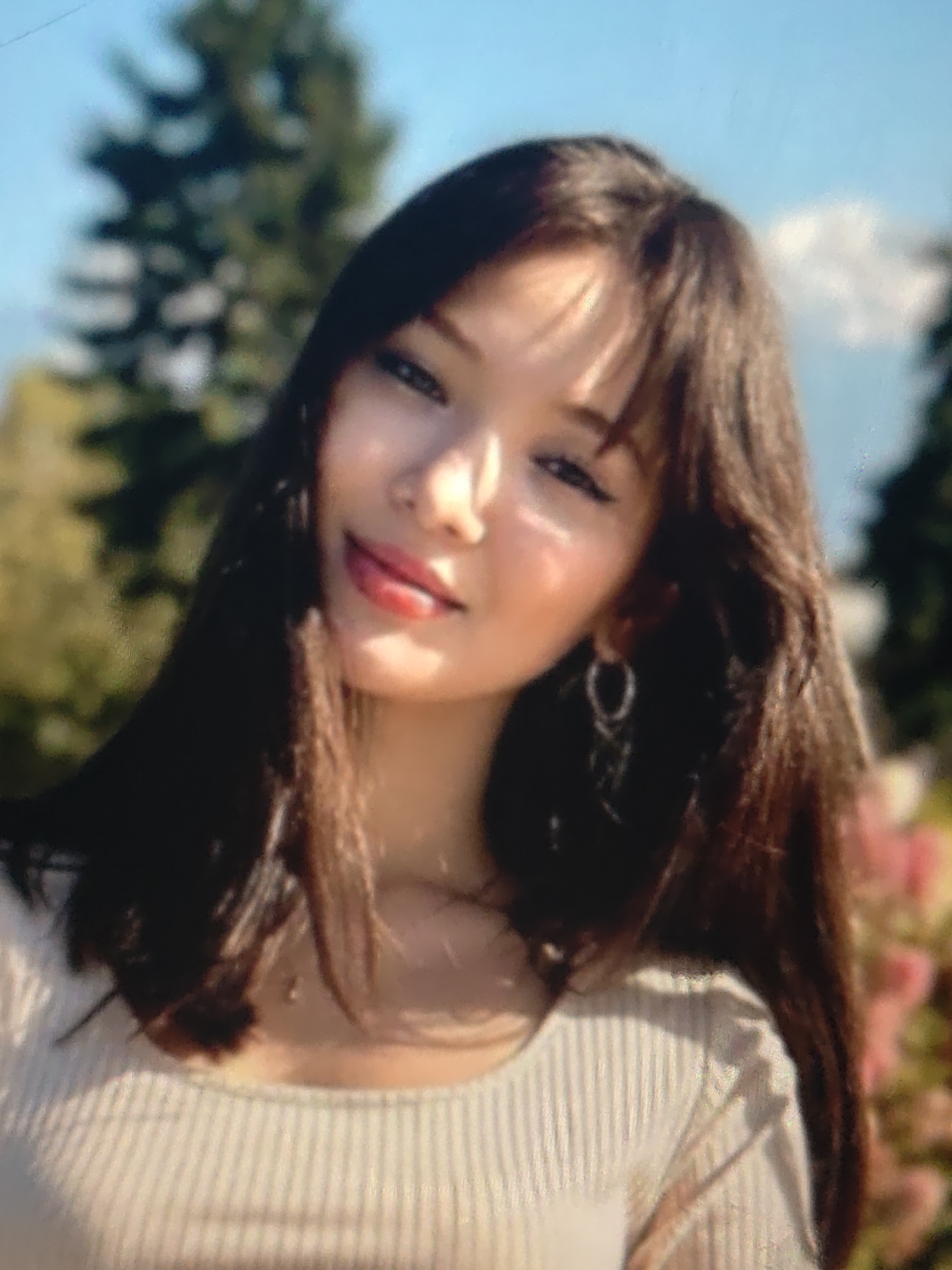 A young woman with long, dark brown hair and bangs is shown from the chest up, outdoors. She is wearing a light-colored, ribbed shirt. She has on a hoop earring in her left ear and appears to be wearing light makeup, including lipstick. The background is blurred, showing trees and foliage under a blue sky. The image is slightly out of focus.
