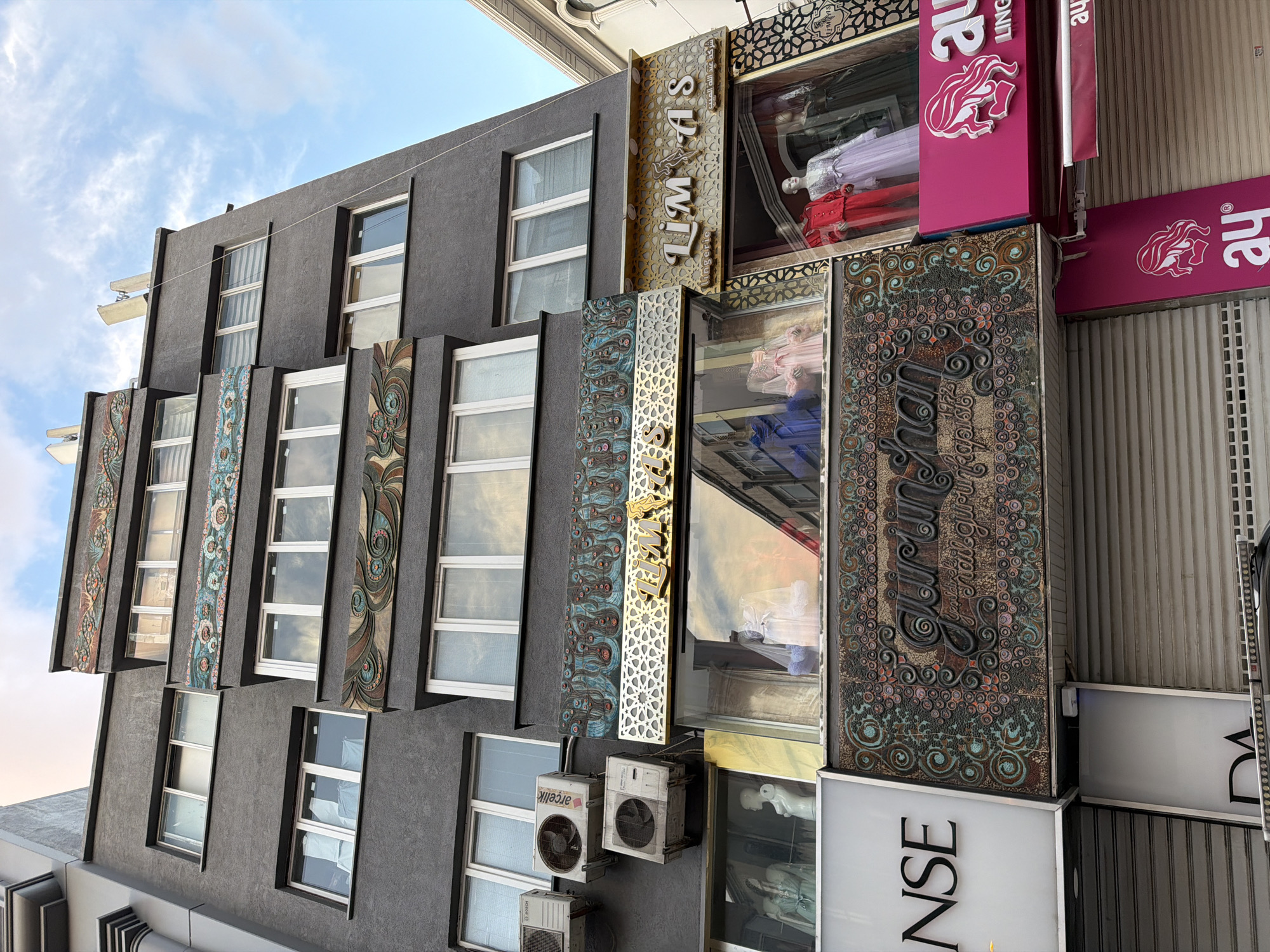 The image shows the facade of a multi-story building in Istanbul, Turkey. The building has a dark gray exterior with several floors of windows. The lower floors feature storefronts with mannequins displaying clothing. The storefronts have illuminated signs. One sign reads "LIMAS lingerie" in a decorative font. Another large, ornate sign reads "Gurunguhan 1972" in a raised, textured font with decorative elements. There are also pink signs with a woman's profile and the text "ay" and "LING" visible. The upper floors have rows of windows, some with blinds. Air conditioning units are mounted on the exterior of the building. The sky above is blue with some clouds. The overall scene depicts a commercial street in Istanbul with a focus on clothing retail.