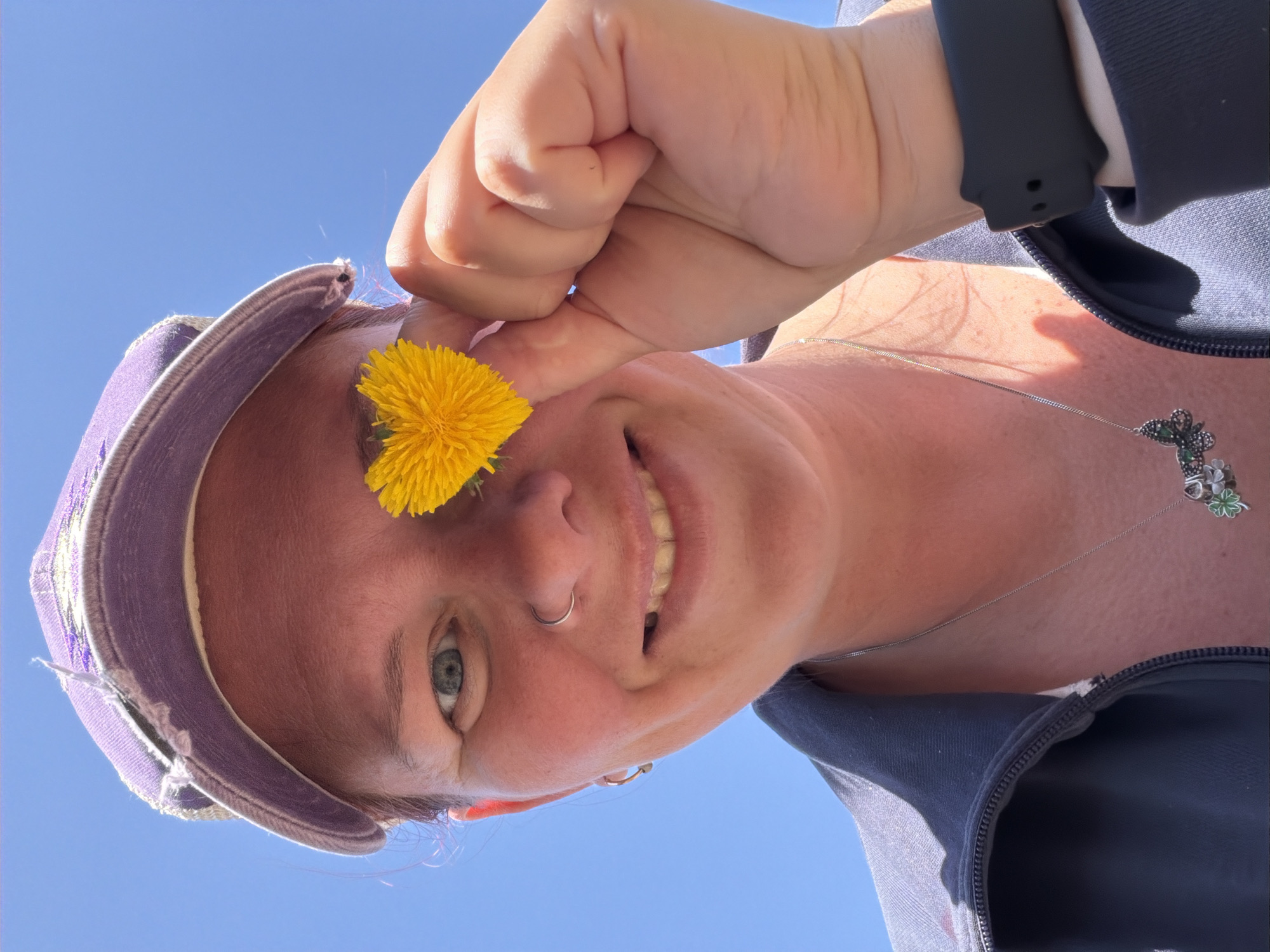 A woman wearing a purple hat and a silver necklace with a butterfly pendant is holding a yellow dandelion flower near her nose. She has a nose ring and is smiling. Her hand is clenched in a fist. She is wearing a dark blue jacket and a black watch. The background is a clear blue sky. There is no information to specify the location in the city of Chaikovsky, Russia.