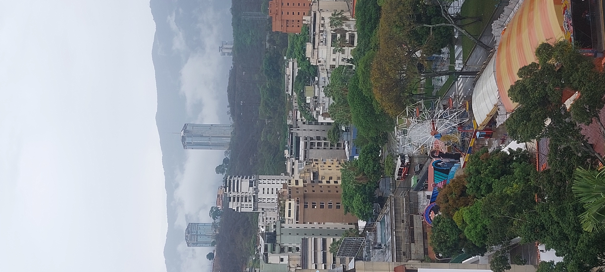 The image depicts a cityscape, presumably in Caracas, Venezuela, featuring a ferris wheel and other amusement park rides in the foreground. A dense collection of buildings of varying heights fills the midground, transitioning into a backdrop of tree-covered mountains obscured by low-hanging clouds and fog. The sky is overcast, suggesting a rainy or misty day. The general conditions appear urban.