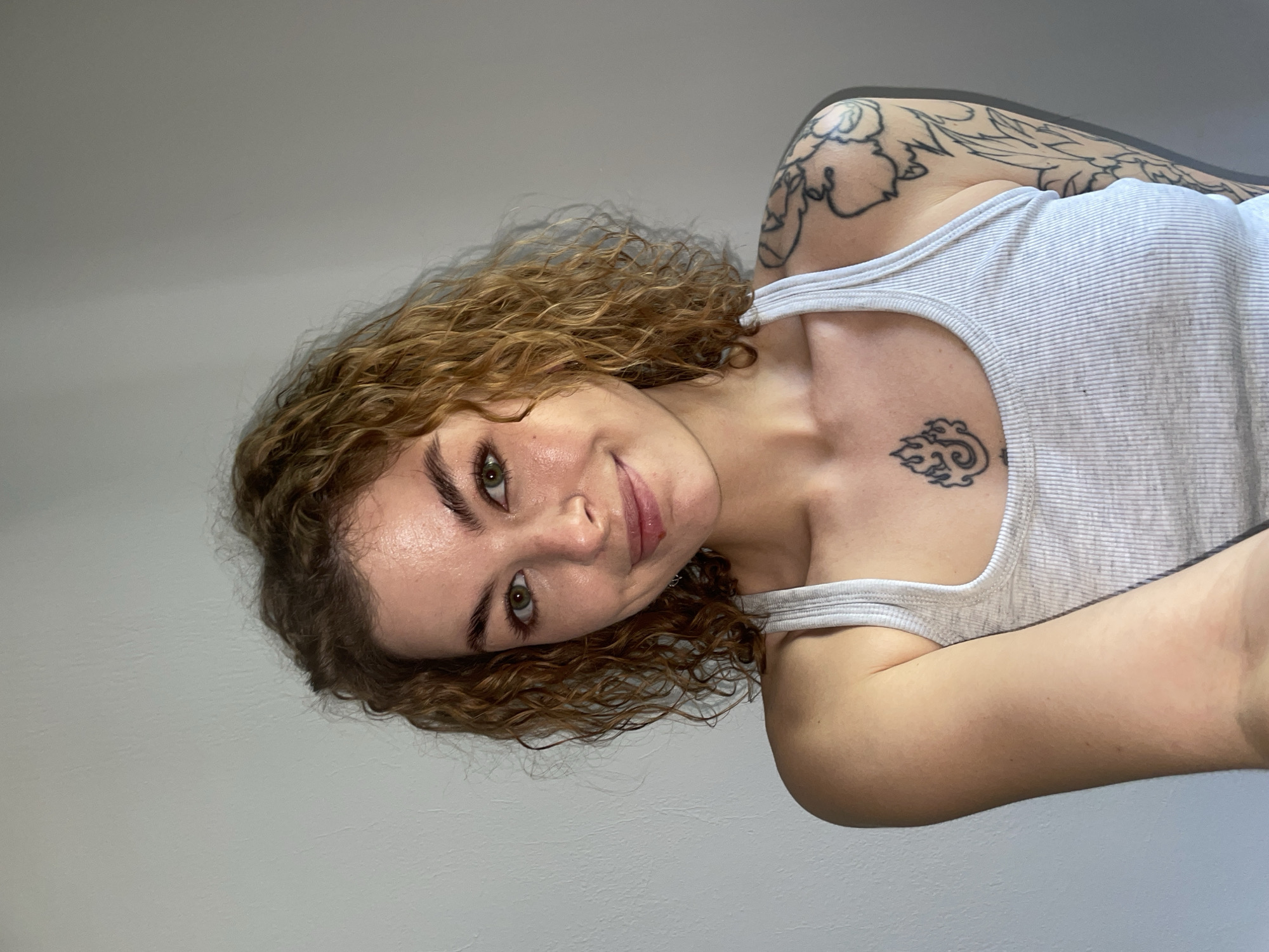 A light-skinned woman with curly brown hair and tattoos is shown. She is wearing a gray tank top. Tattoos are visible on her upper left arm and chest. The background is a plain gray wall. There is no evidence to suggest that the image was taken in Boreti, Montenegro.