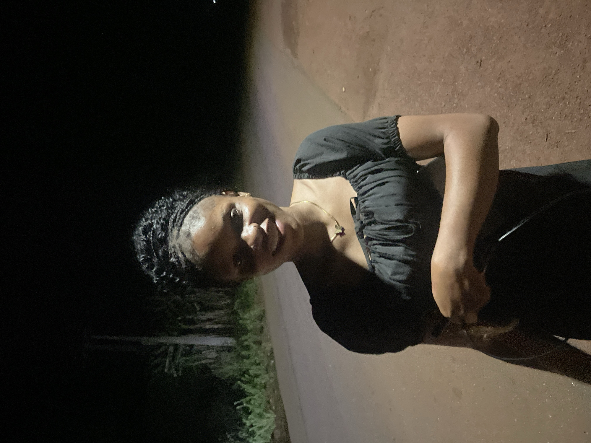 A woman with dark hair braided close to her head stands on a dirt road in Ogbe, Nigeria. She wears a black top and a gold necklace. The background is dark, with vegetation visible on one side of the road. The lighting suggests it is night.
