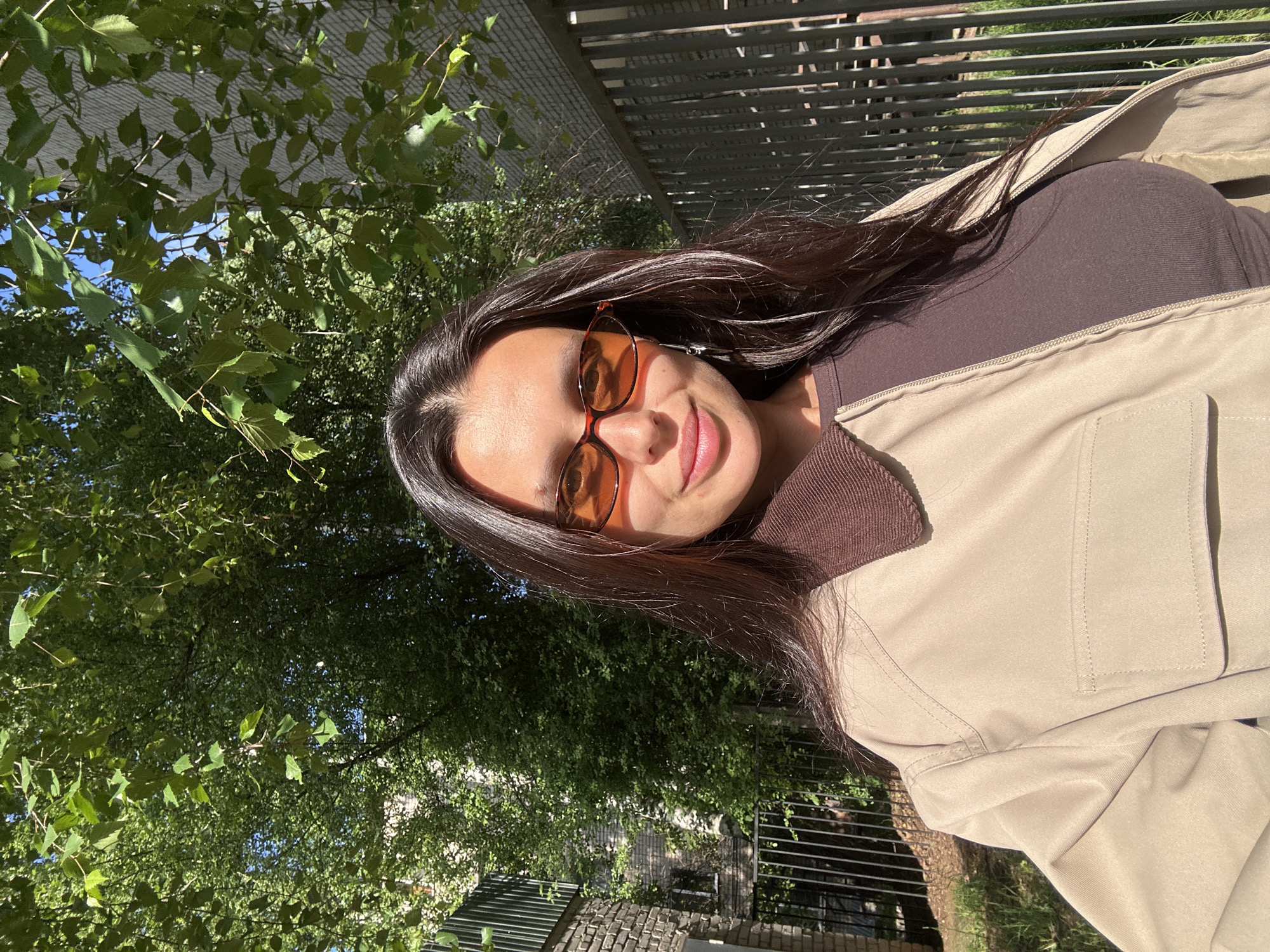 The image, rotated 90 degrees counter-clockwise, features a young woman smiling and posing outdoors on a bright, sunny day in Chaikovsky, Russia. She has long, dark brown hair and is wearing sunglasses with prominent orange-tinted lenses. Her attire includes a dark brown top and a light beige jacket with a matching brown collar. The sunlight creates strong contrasts, highlighting her features and clothing.
The background is rich with dense green foliage from trees, with patches of bright blue sky visible through the leaves, suggesting a natural outdoor environment like a park or garden. A dark, horizontal metal fence or trellis structure runs across the upper right portion of the scene. Below the vibrant greenery, elements of an urban landscape, such as brickwork and additional fencing, can be seen, indicating a blend of natural and built surroundings. The woman's expression is pleasant and appears happy. There is no visible text.