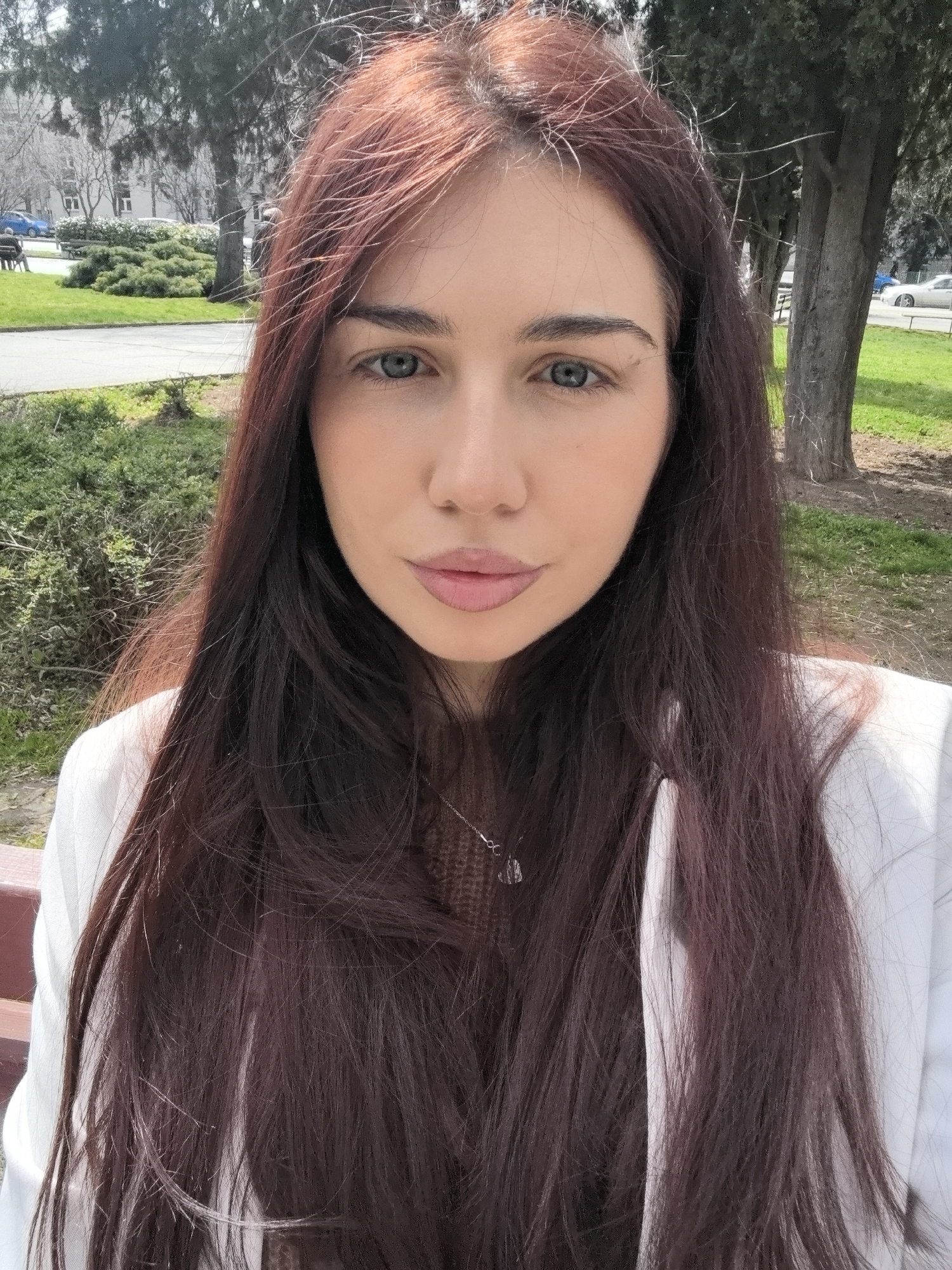 A medium shot captures a woman outdoors in Stara Zagora, Bulgaria. The woman, with long, auburn hair and light-colored eyes, is wearing a white blazer over a brown top and a necklace. She is positioned on what appears to be a park bench, with a park and buildings visible in the background. A tree is also visible behind her on the right side of the image. The setting suggests daytime with natural light.