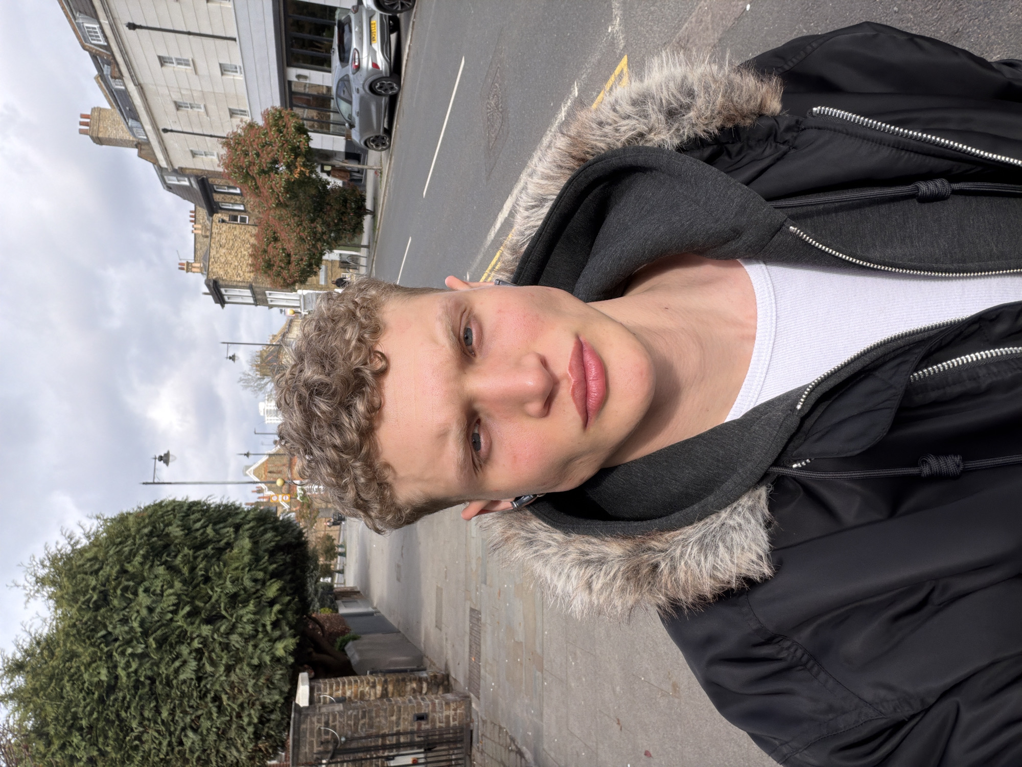 A young man with light complexion and short, curly hair is the primary subject of the photo. He is wearing a white undershirt, a gray hooded sweatshirt, and a black jacket with fur trim around the hood. He is standing on a sidewalk next to a road with a car parked in the distance. Behind him are buildings characteristic of London, including a multi-story building with white exterior walls and a building with brick and stone construction. A small tree with reddish leaves is also visible. The sidewalk is made of concrete slabs, and a large, dense bush is on the left side of the frame. The photo is taken outdoors during daylight with natural lighting.