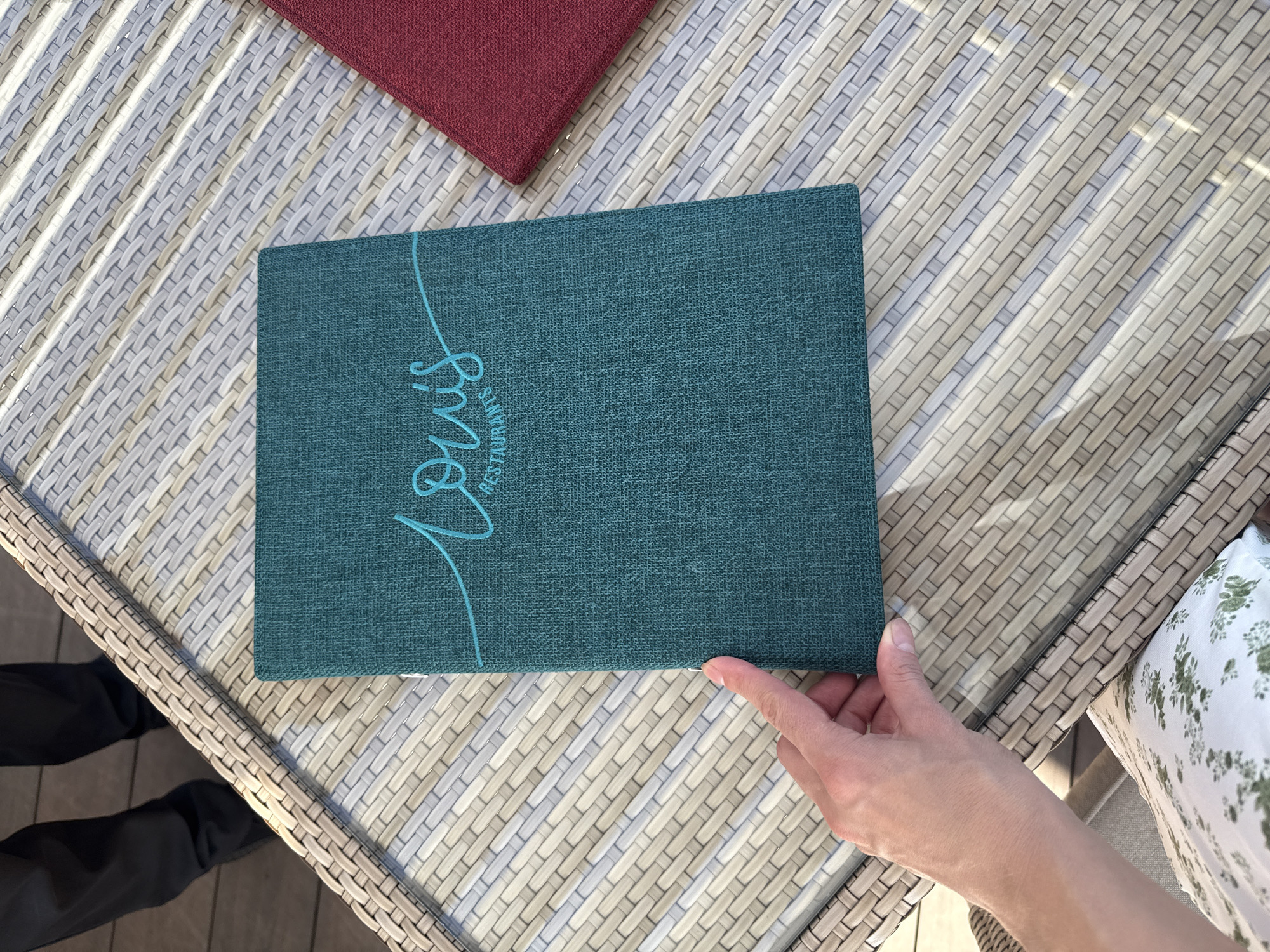 A person's left hand is seen in the bottom left corner of the image, holding open a teal-colored menu with "lou's RESTAURANTS" written in cursive on the cover in a lighter teal color. The menu is lying on a woven light-gray table, with a red menu or placemat partially visible to the right. In the background, a person wearing dark pants is standing. The scene appears to be outdoors, with natural light casting shadows on the table. The location is the city of Kazan, Russia.