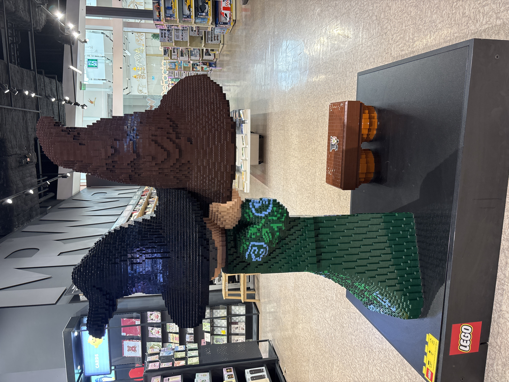 This image depicts a large LEGO sculpture of two overlapping wizard hats, one black and one brown, appearing to be made from LEGO bricks. The hats are perched atop a green LEGO structure that resembles a serpent. Below the sculpture, a black platform displays a LEGO logo on the left side and a small LEGO chest on the right. The sculpture is situated indoors, within a retail environment that appears to be a store. Shelves stocked with merchandise are visible in the background, along with signage that includes the word "DeFacto". The floor is tiled with a light-colored pattern. The lighting suggests it is daytime. There are no people visible in the image. The location in the city of Astana, Kazakhstan, is not discernible from the visual information provided in the image.