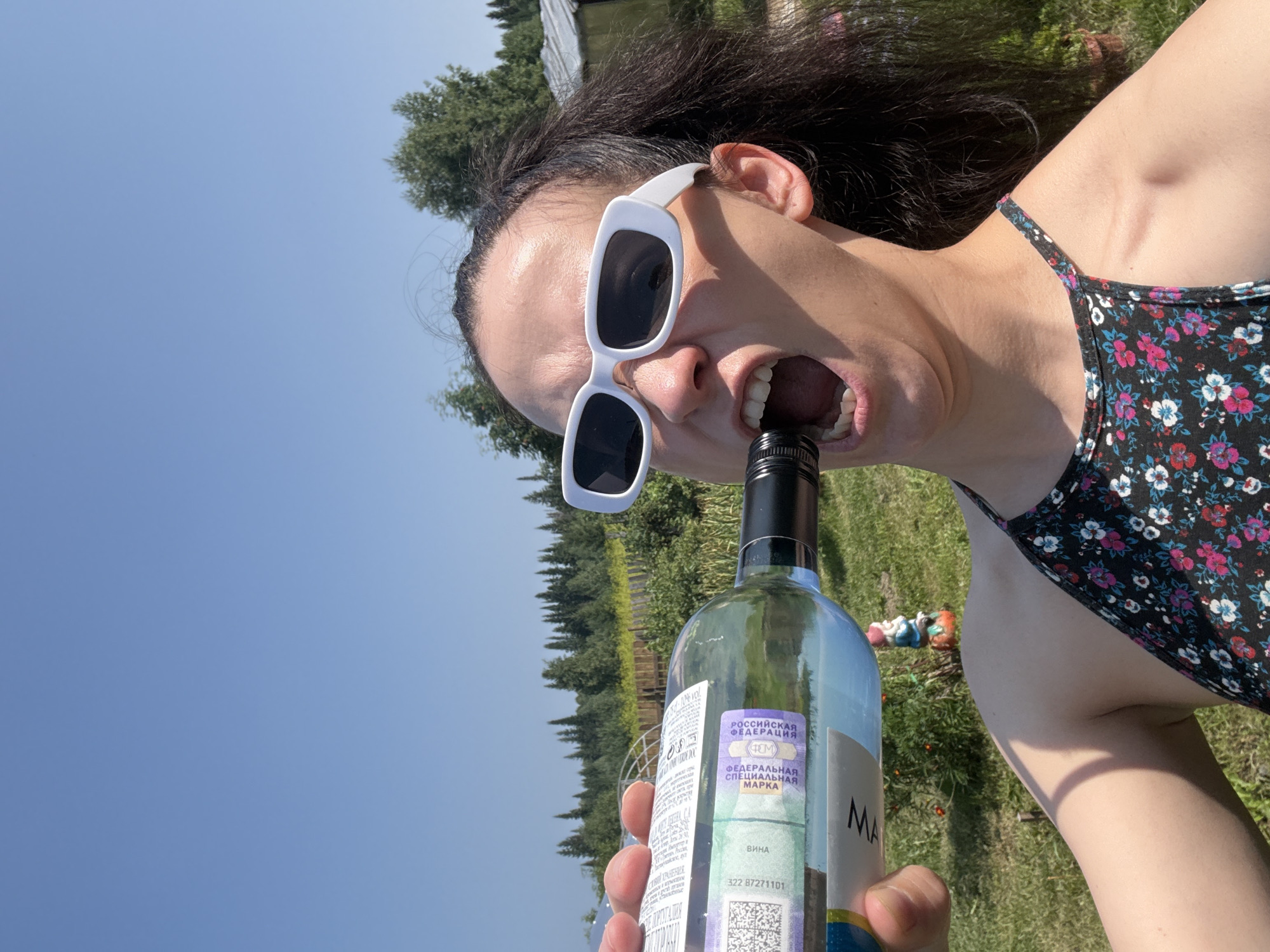 A woman is holding a bottle of white wine with her left hand, bringing the neck of the bottle towards her open mouth. She is wearing white rectangular sunglasses and a floral-patterned top. The background shows a clear blue sky, trees, and a small garden gnome. The scene appears to be outdoors in a rural or suburban setting, possibly in Zasechnyi, Russia, given the presence of Russian text on the wine label, which indicates it is a special mark for wine. The woman's facial expression suggests she is about to drink from the bottle.