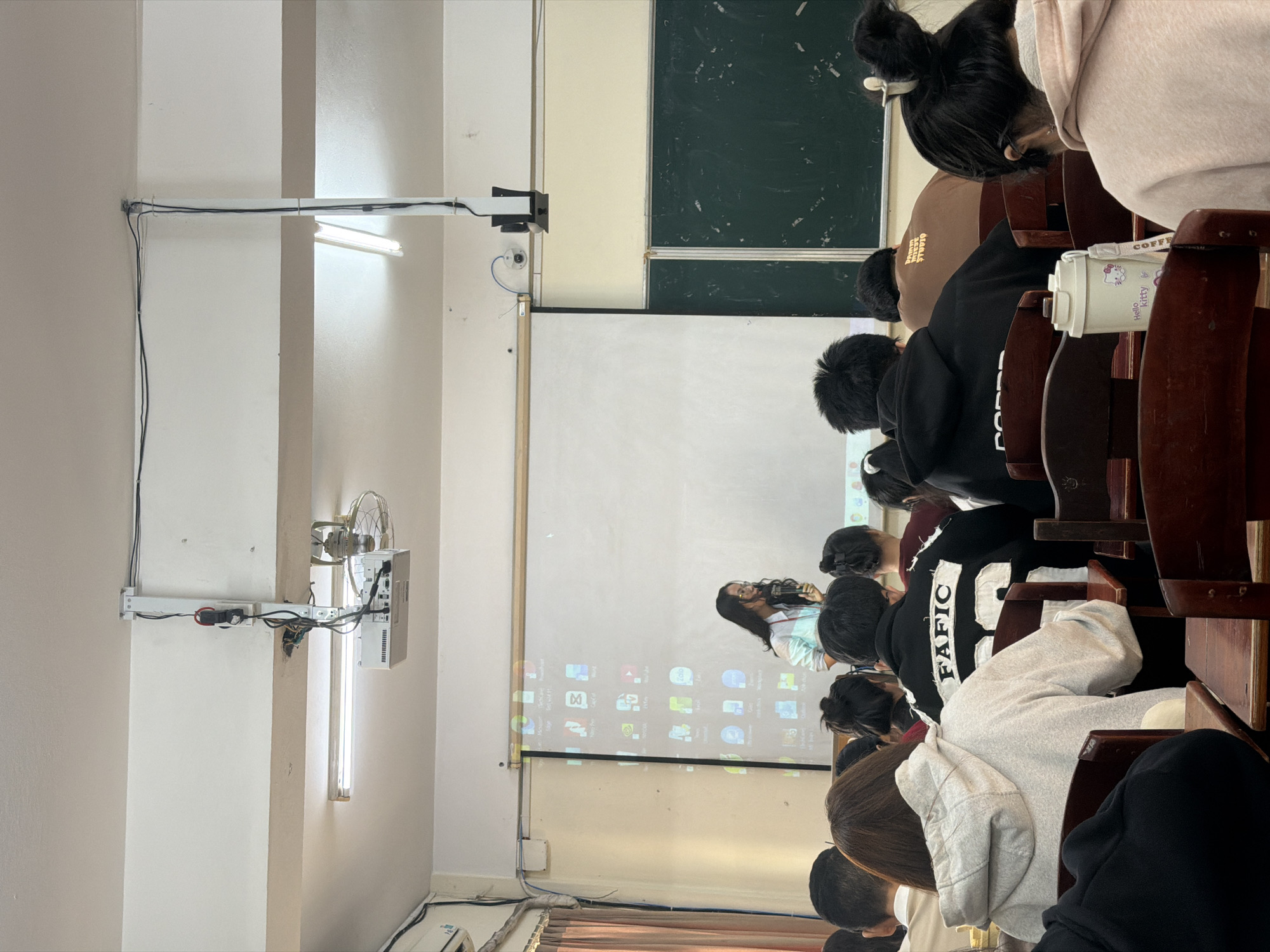 The image captures a classroom in Đà Nẵng, Vietnam, during a lecture or presentation. The perspective is from the rear of the room, looking towards the front where students are seated in rows at wooden desks and chairs. Most students are seen from behind, with dark hair, some tied up. A teacher or presenter stands near a large white projection screen, which displays a computer desktop with various application icons.
Above the projection screen, a dark green chalkboard is visible, with a white writing board positioned beneath it. The classroom's functional setup includes multiple fluorescent light fixtures mounted on the ceiling and side walls, accompanied by exposed electrical wiring. A small, old-fashioned metal fan is mounted high on the right-hand wall, indicating a means of air circulation. A projector is secured to a wall-mounted apparatus, casting the digital display onto the screen.
Among the students' belongings, a white thermos with "Hello Kitty" branding and a pink bow is visible on a desk. Text partially visible on student clothing includes "FANNO STUDIO" on a light brown hoodie and "FAFICA" (or similar characters) on a black jacket. The lighting in the room, supplemented by artificial lights, suggests it is daytime.