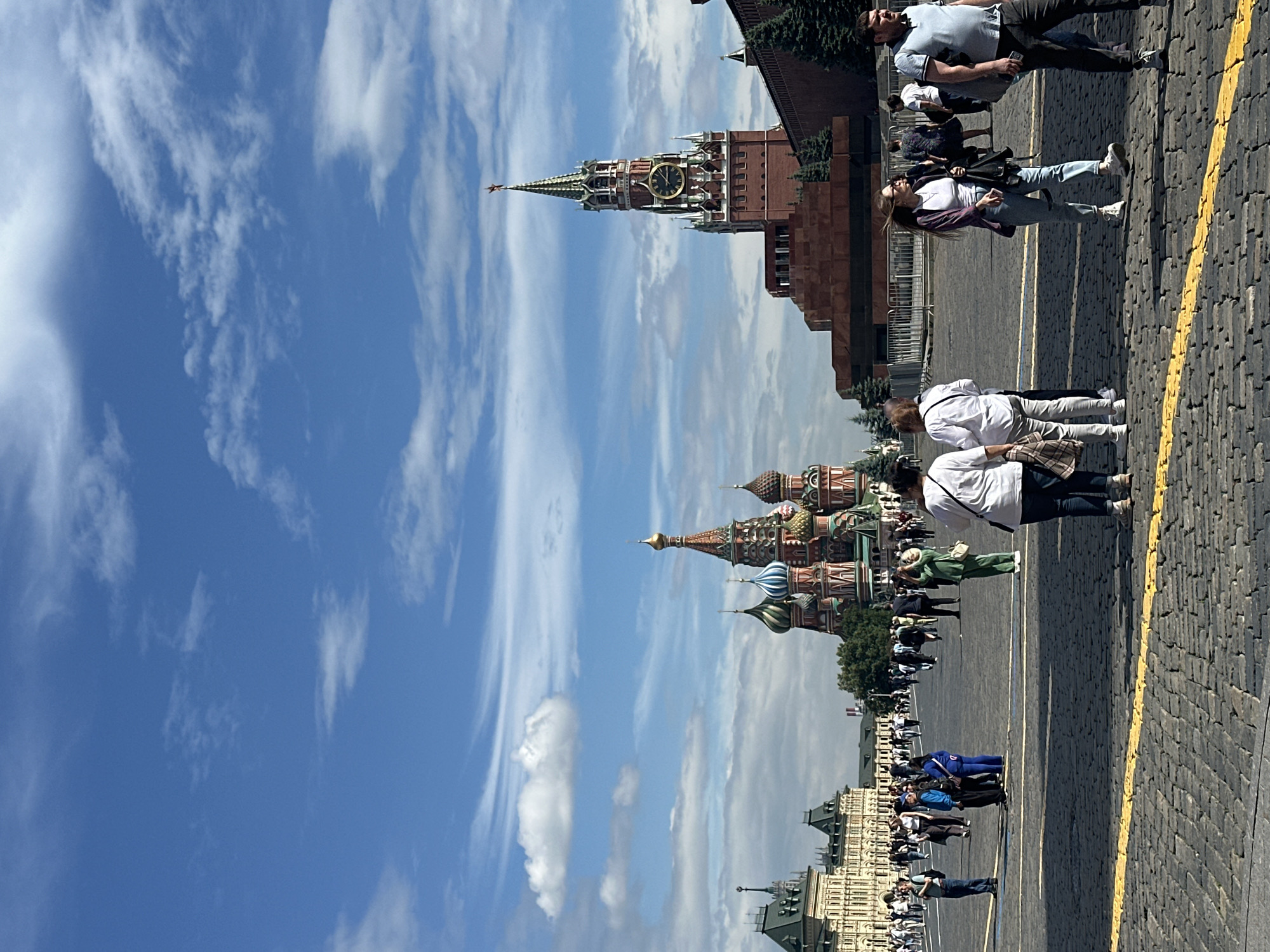 The image shows a panoramic view of Red Square in Moscow, Russia, on a clear day with scattered clouds. In the background, Saint Basil's Cathedral, with its iconic onion domes, is prominently featured on the left. To the right of the cathedral, the Spasskaya Tower, part of the Kremlin wall, stands tall with a clock face visible. In the foreground, a cobblestone plaza is populated by a number of people walking and standing around. Some individuals are captured mid-stride, while others are engaged in conversation or observing the surroundings. The overall scene depicts a bustling tourist area with historical landmarks.