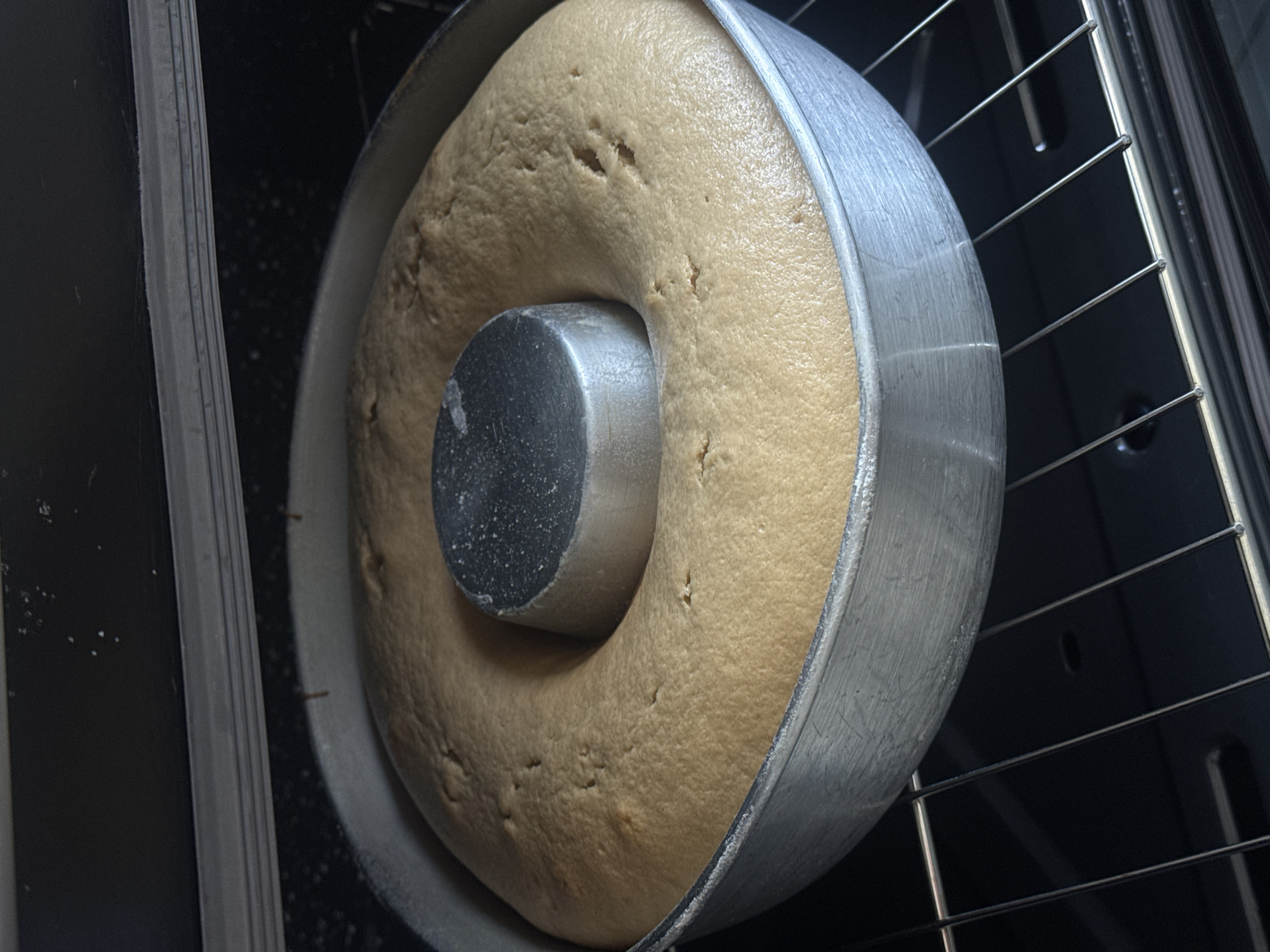 A close-up photograph shows a cake baking in a bundt pan inside an oven. The cake is golden brown and has risen to the top of the pan. The bundt pan is made of silver metal and has a central tube. The oven rack is visible below the pan, with the oven's interior appearing dark. The location is not discernible from the given image.