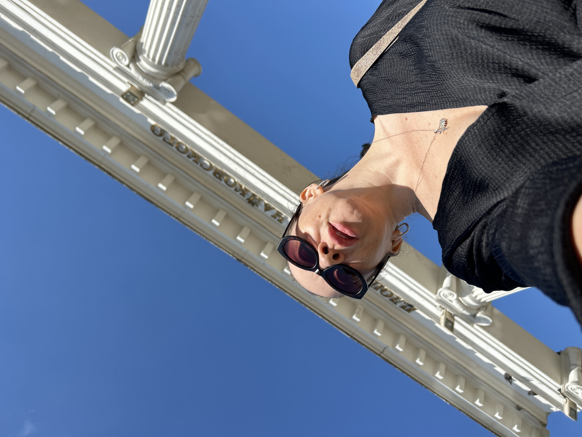 A low-angle, outdoor, daylight, selfie-style photograph shows a bald woman wearing black clothing and sunglasses. She is looking up and to the right, her mouth slightly open. She is wearing a silver necklace with a small pendant and a ring in her left ear. Behind her, a white architectural structure with columns and decorative molding is visible against a clear blue sky. Gold lettering on the structure reads "ЧАЙКОВСКОГО" which translates to "Chaikovskogo," indicating the location is likely in the city of Chaikovsky, Russia, or named after Pyotr Ilyich Chaikovsky, the famous Russian composer. The woman is wearing a light-colored strap over her right shoulder.