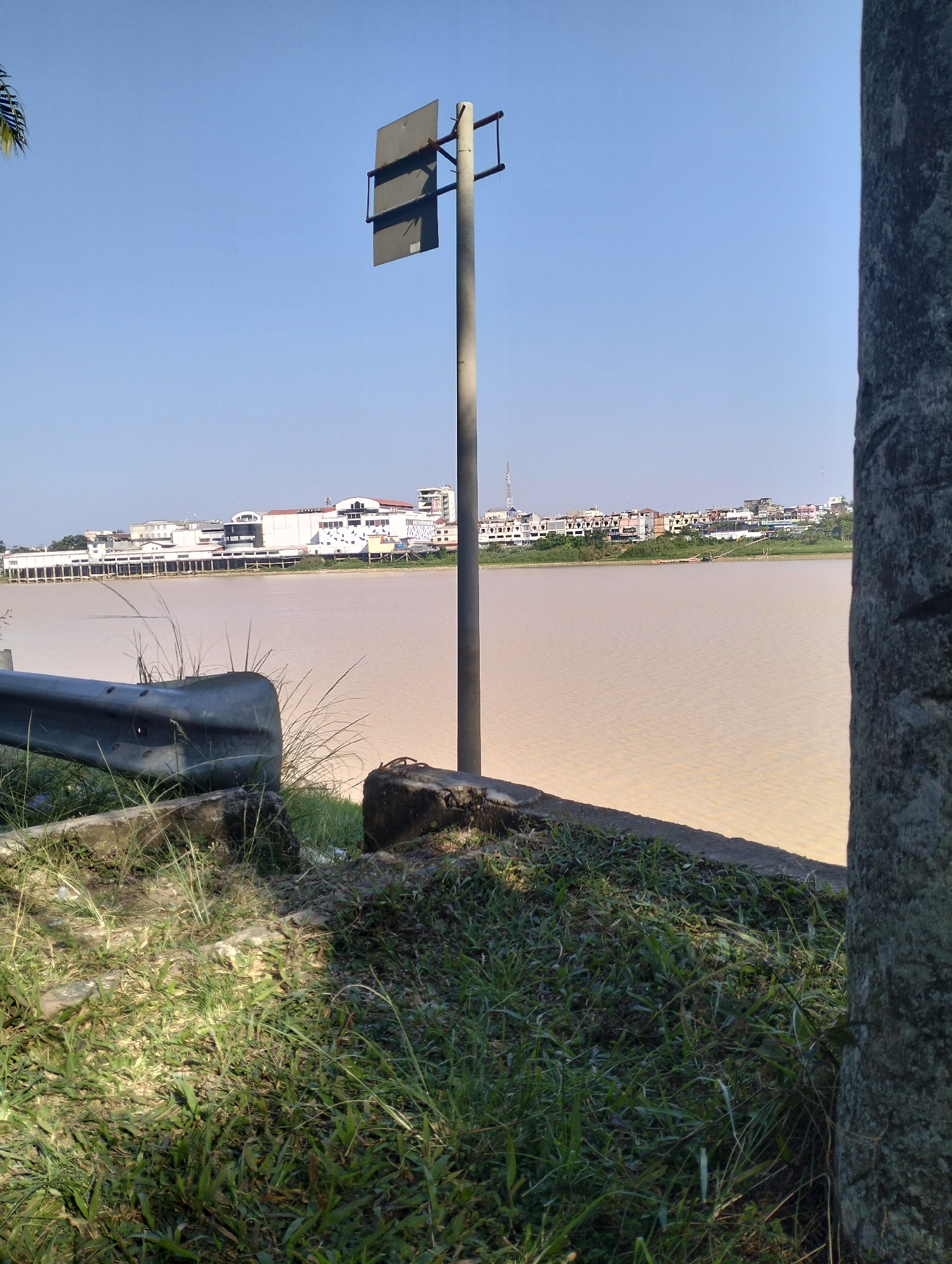 A wide, muddy river flows through a city in Jambi, Indonesia, under a clear blue sky. The foreground of the image is dominated by lush green grass and a section of a metal guardrail. A tall, slender concrete pole with a dark rectangular sign attached to it stands in the middle ground, casting a faint shadow. Beyond the river, a cityscape unfolds, characterized by a collection of low-rise buildings with white facades and diverse architectural styles, suggesting a mix of commercial and residential structures. The buildings are densely packed along the riverbank, with some taller structures visible in the distance. The river itself appears calm, with a few ripples on its surface. The overall impression is of a sunny day in an urban environment situated along a waterway.