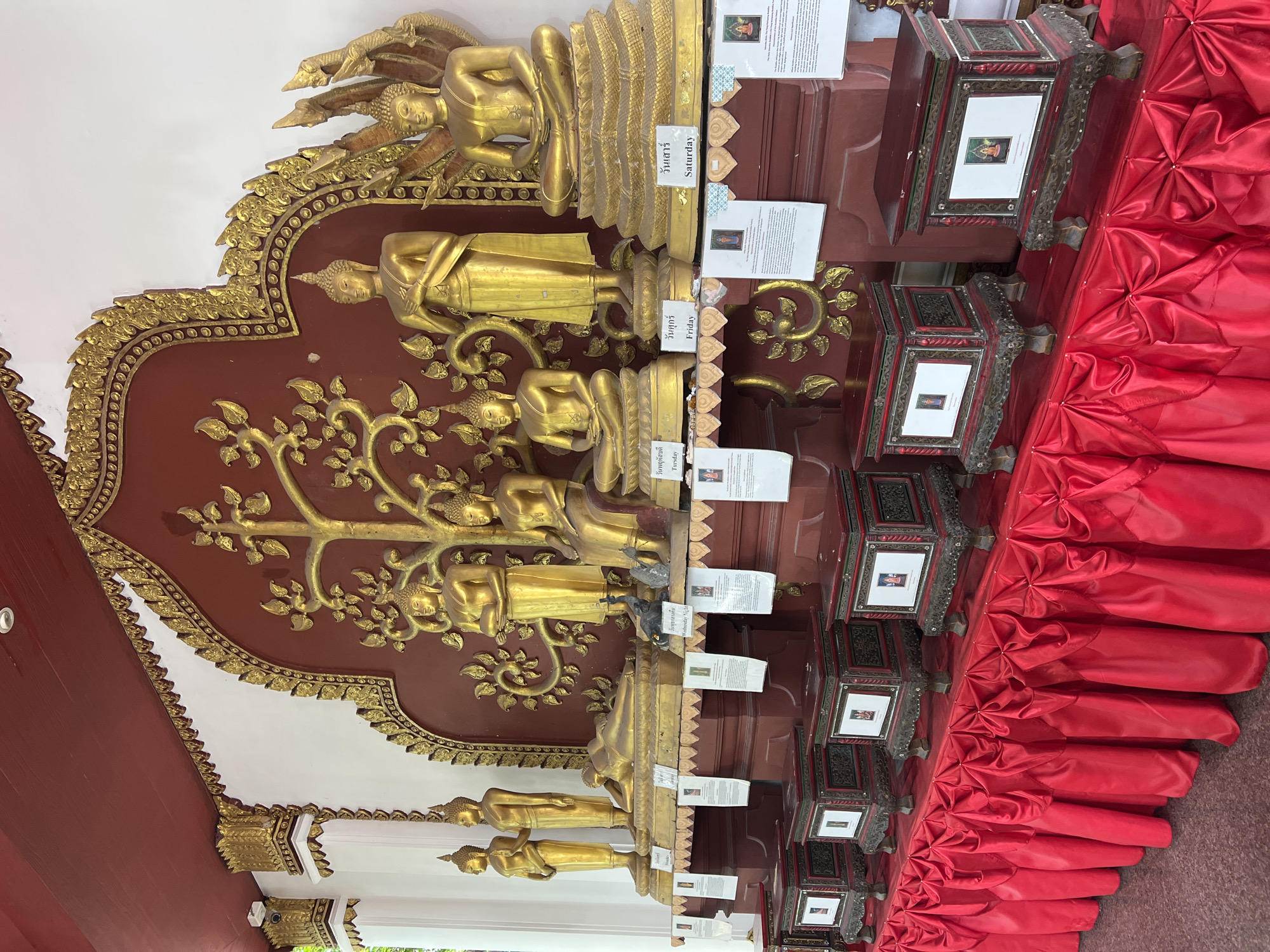 This image shows a display of golden Buddha statues in the city of Baan Hua Thanon, Thailand. The statues are arranged on a raised platform against a red backdrop adorned with intricate gold carvings, including a tree of life motif. Several statues depict Buddha in various poses, such as standing, reclining, and seated. In front of the statues are small, ornate wooden boxes, likely for donations or offerings. These boxes are covered by a red draped fabric. White informational cards are placed in front of some of the statues and boxes. The overall scene is a religious and cultural display, likely within a temple or shrine.