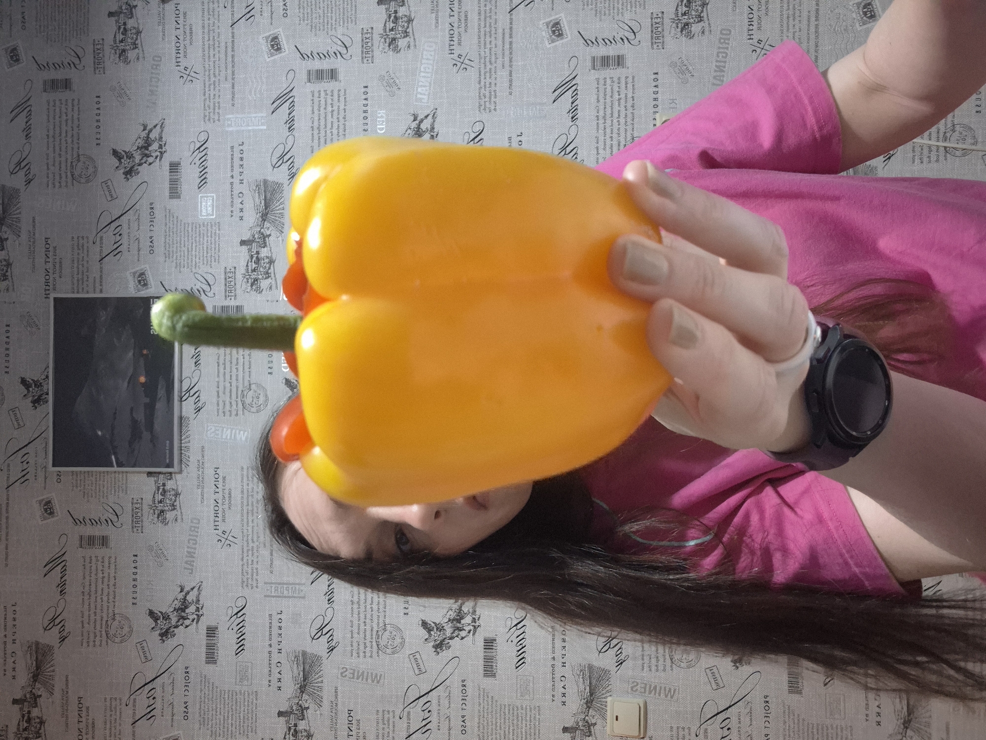 A woman wearing a pink shirt and a black smartwatch holds a large yellow bell pepper in front of her face. The background features a wall with a newspaper print design. A framed picture is also visible on the wall.