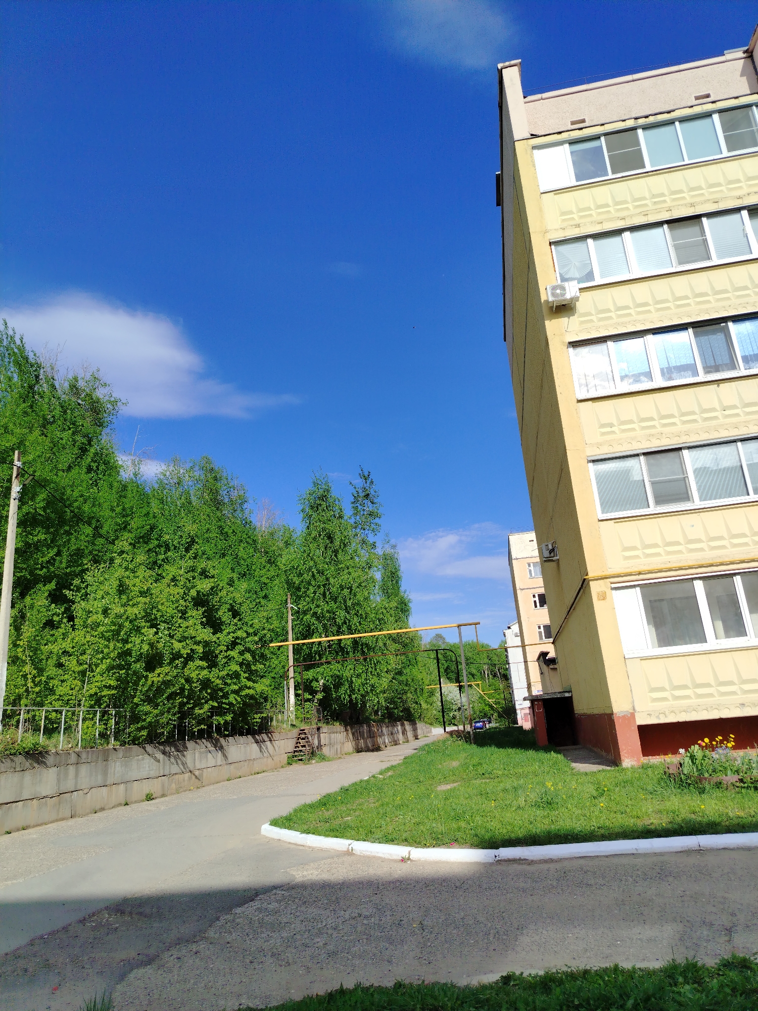 The image depicts a street scene in Noviy Urengoy, Russia. The sky is a clear blue with a few scattered clouds. A tall, light-yellow apartment building dominates the right side of the frame, featuring multiple floors with windows and a few visible air conditioning units. A smaller building is visible behind the taller one.
To the left of the building is a paved street, partially shaded, with a concrete barrier alongside it. A dense area of green trees and foliage lines the other side of the street. Yellow pipes and some metallic structures can be seen near the trees. A grassy area with some yellow flowers lies between the building and the street.