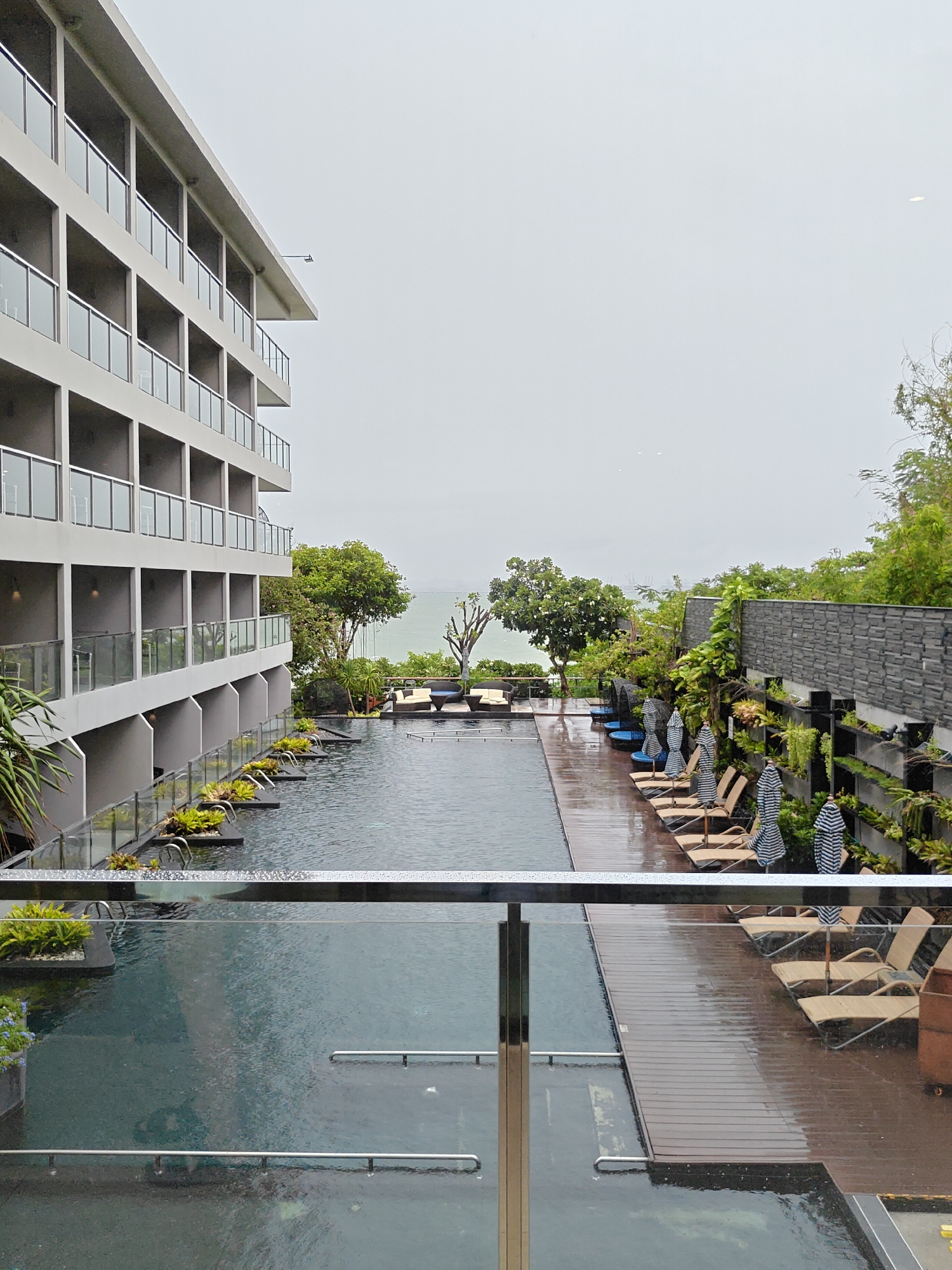 This image depicts a serene resort swimming pool area in Pattaya City, Thailand, on an overcast day. The resort building, characterized by its modern, multi-story architecture with balconies and glass railings, dominates the left side of the frame. The swimming pool is a prominent feature, with its dark water reflecting the muted sky and surrounded by a wet wooden deck. Lounge chairs with striped umbrellas are neatly arranged on the deck, some partially closed, suggesting a break from activity. Lush green vegetation, including trees and potted plants, adds a touch of nature to the scene. In the background, the sea is visible, meeting the hazy horizon under a gray sky. The overall ambiance is tranquil and suggests a peaceful, possibly rainy, day at a tropical resort.