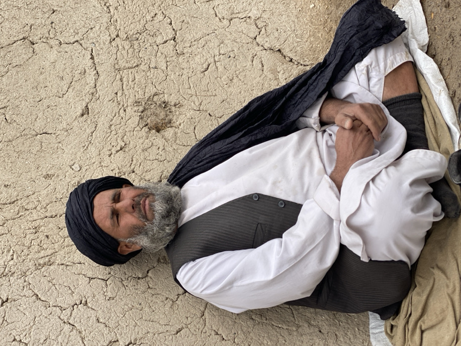 An elderly man with a grey beard lies on his side, possibly sleeping. He is wearing a black turban and vest over a white shirt. He has grey pants on and there is a black cloth draped around his upper body. His hands are clasped in front of him. He is lying on a cracked, light brown surface, possibly a dirt or mud floor, with a lighter colored cloth visible near his feet.