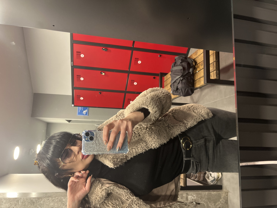 A mirror selfie captures a female individual in what appears to be a changing room or locker room, likely within a fitness facility. The precise location within Varna, Bulgaria, cannot be determined from the visual evidence.
The subject, viewed from the waist up, wears a black ribbed turtleneck, a light brown faux-shearling jacket, dark grey jeans, and a black belt with a gold-colored interlocking circle buckle. Her dark hair features bangs and is partially secured with a hair claw. She wears large, dark-tinted sunglasses, dark eyeliner, and reddish lipstick. Gold earrings are partially visible. She holds a smartphone with a glittery silver case in her right hand; her nails are polished in light blue/silver glitter and nude/brown tones.
The background shows rows of red metal lockers with black frames and circular ventilation holes, positioned above wooden benches with dark metal supports. A black duffel bag rests on a bench to the right. The walls are light grey, and a blue and white informational sign is visible on the left wall. Recessed ceiling lights are present. A pair of light-colored sneakers is partially reflected on the floor below a bench on the left.