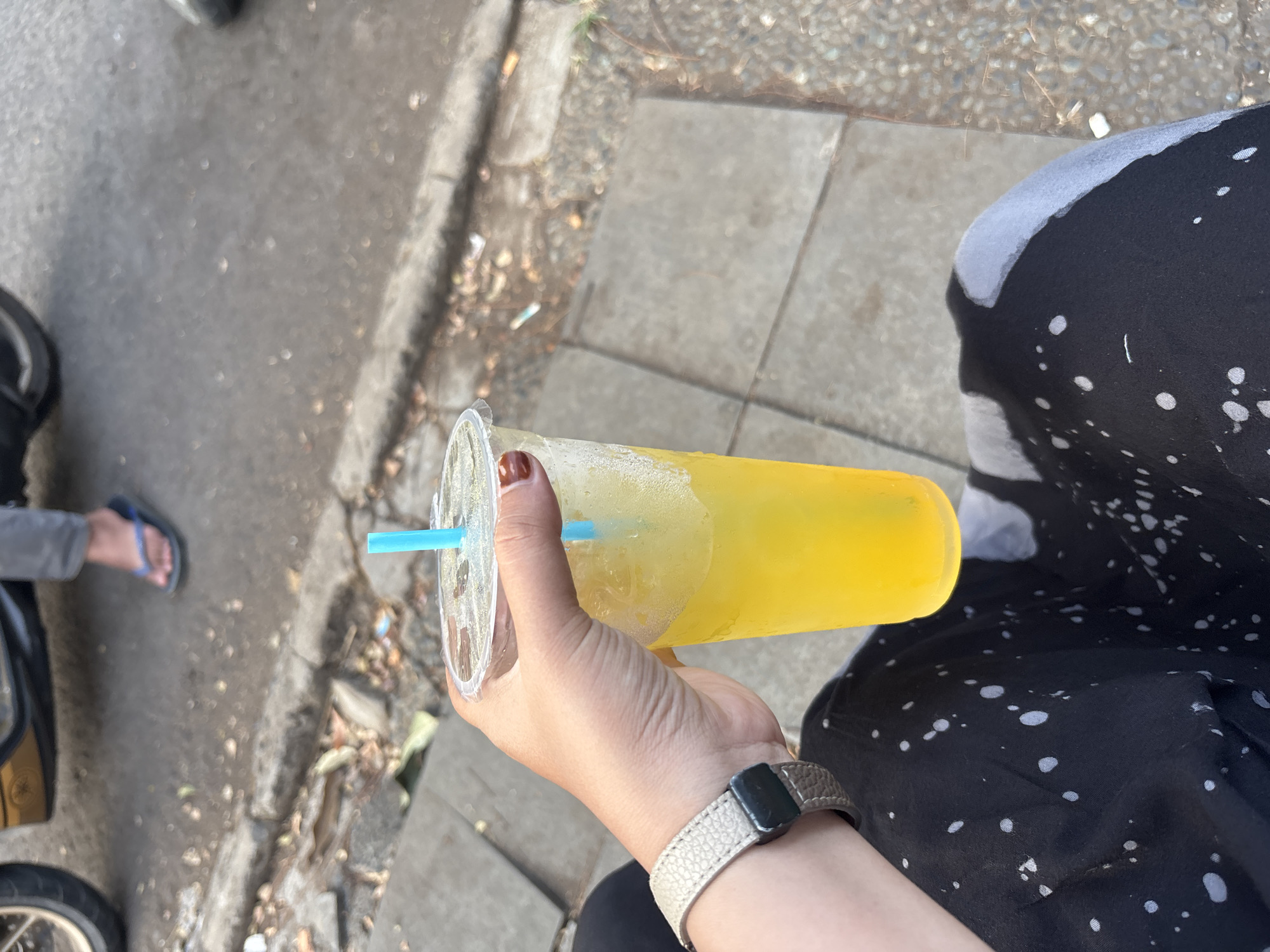 A person's hand, adorned with a watch and painted nails, holds a clear plastic cup filled with a yellow beverage and ice, topped with a blue straw. The cup is positioned in the foreground, obscuring the lower half of the body which is dressed in black patterned clothing. In the background, a person's legs wearing gray pants and blue flip-flops are visible, with a portion of a motorcycle behind them. The scene appears to be outdoors on a paved surface, possibly a sidewalk, with debris scattered on the ground, suggesting an urban environment in Cirebon, Indonesia.