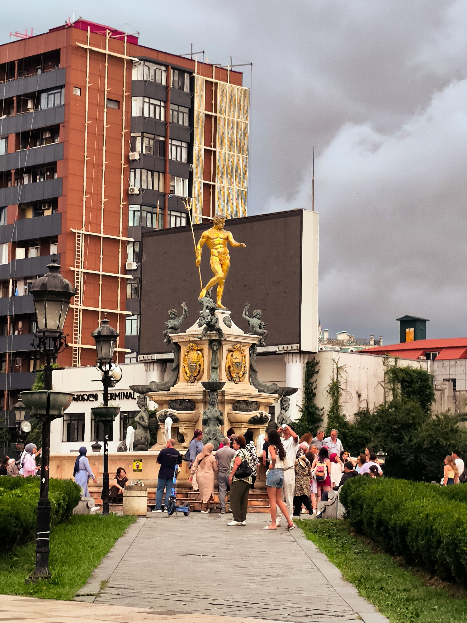 The image depicts a public square in Batumi, Georgia, featuring the iconic statue of Neptune. In the foreground, a pathway is lined with lush green grass and hedges. Ornate lampposts stand to the left, casting a vintage ambiance. A crowd of diverse individuals is gathered around the Neptune statue, which is prominently positioned atop a multi-tiered fountain. The statue, cast in gold, depicts Neptune holding a trident. The fountain itself is adorned with intricate sculptures and ornate details. Behind the fountain, a modern building with a reddish-orange facade and dark grey window panels rises, contrasting with the classical architecture of the fountain. A large, dark billboard is affixed to this building. Further in the background, glimpses of other buildings and lush green trees add depth to the urban landscape. The sky is overcast with dramatic, dark clouds, suggesting an impending storm or recent rainfall. People are engaged in various activities: some are posing for photographs, others are conversing, and a few are observing the fountain. The overall scene captures a lively moment in a bustling city square.