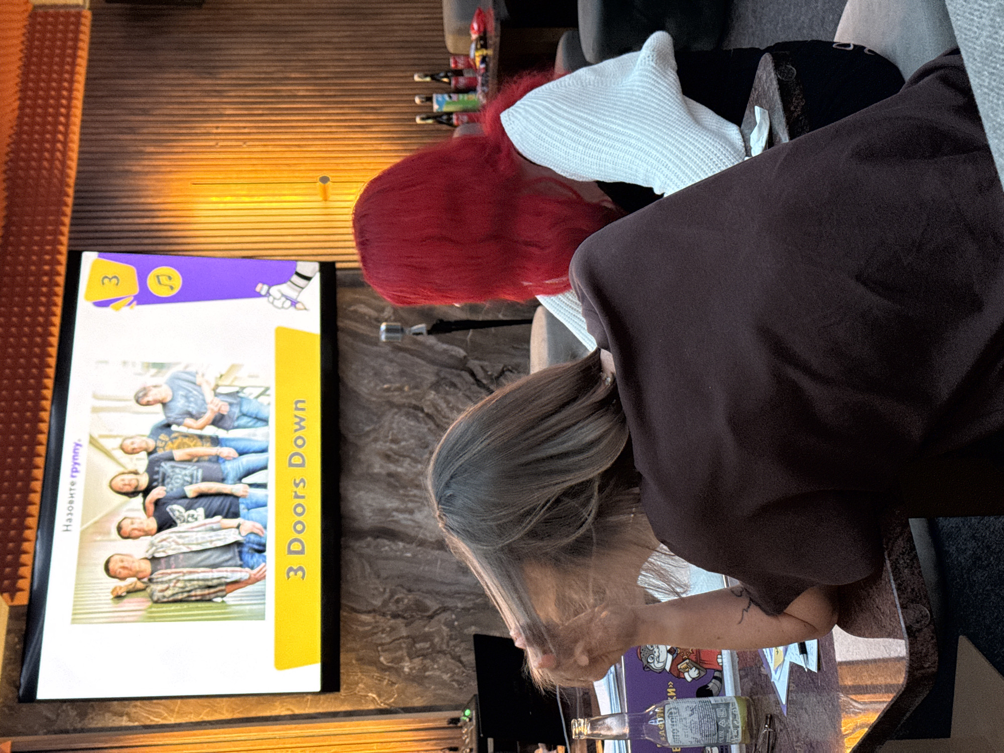 Inside a bar or entertainment venue in Budva, Montenegro, two women are engaged with a television screen displaying a quiz game. One woman has red hair and wears a white sweater, while the other has long, light brown hair and a dark brown shirt. The television screen features a photo of the band "3 Doors Down" as part of the quiz. The venue has textured walls and warm lighting. Beverages are present on the table in front of the women.
