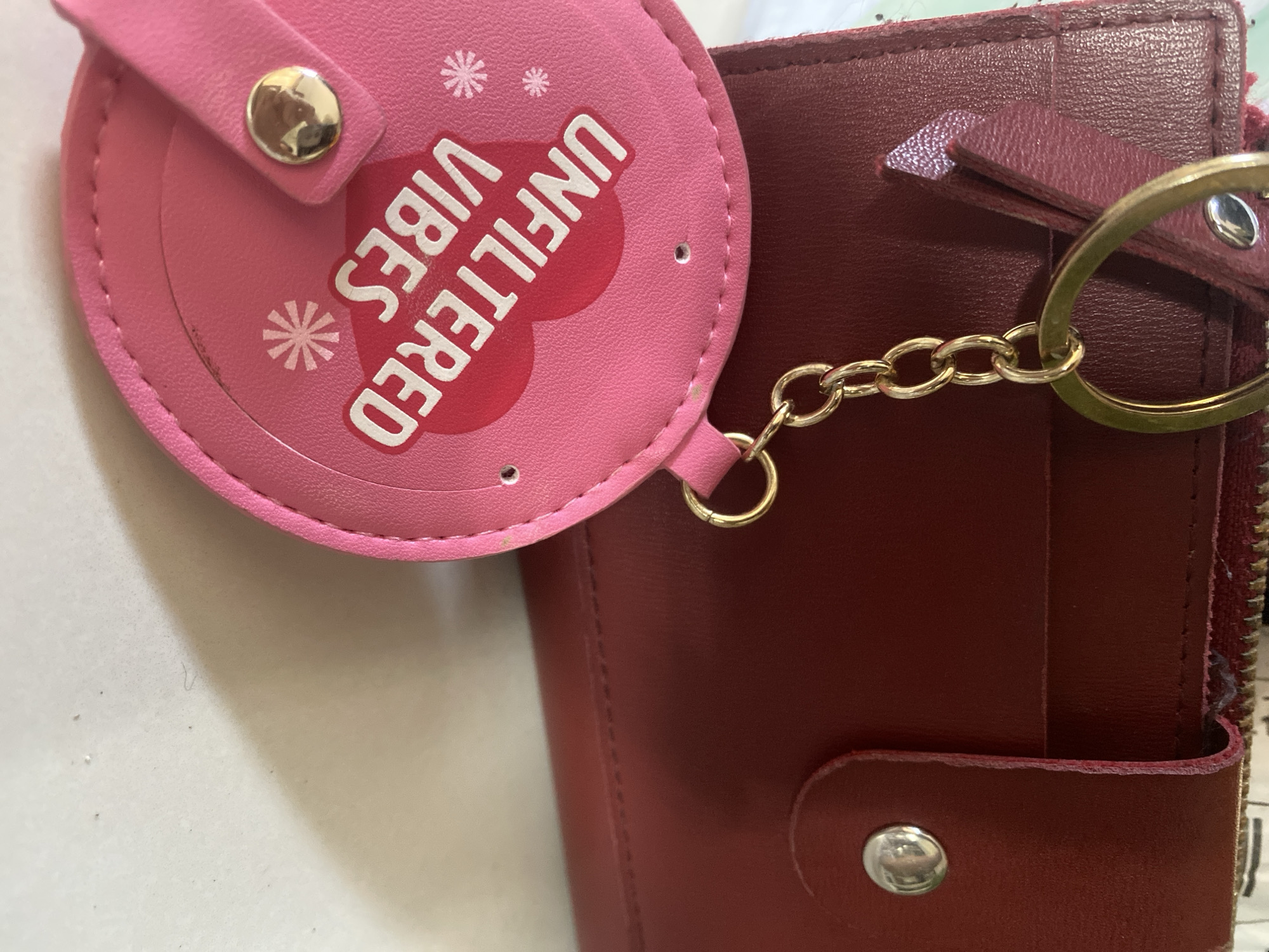 The image presents a close-up view of a pink, circular keychain attached to a red wallet. The keychain has the words "UNFILTERED VIBES" printed in white on a pink background. It also features a metal button and decorative asterisk symbols. The red wallet is made of a leather-like material with a button closure and gold-colored zipper. A gold-colored chain connects the keychain to a loop on the wallet. The background is a white surface with some small specks of dirt. There is no information available about this being taken in Davao City, Philippines.