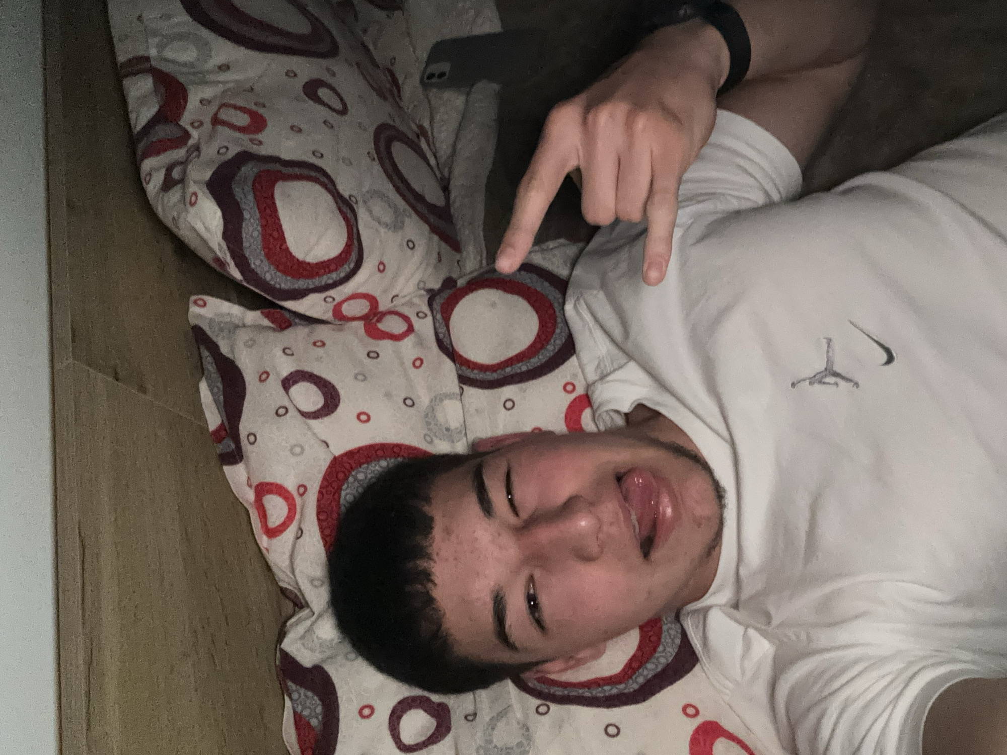 A young man with dark hair lies on a bed covered in a patterned duvet and pillow. He wears a white T-shirt with a logo. He is sticking out his tongue and making the "rock on" hand gesture. A smart watch is visible on his left wrist. A smartphone is also on the pillow next to his head. The location is not discernible in this image. There is no data confirming that the image was taken in the city of Zlatograd, Bulgaria.