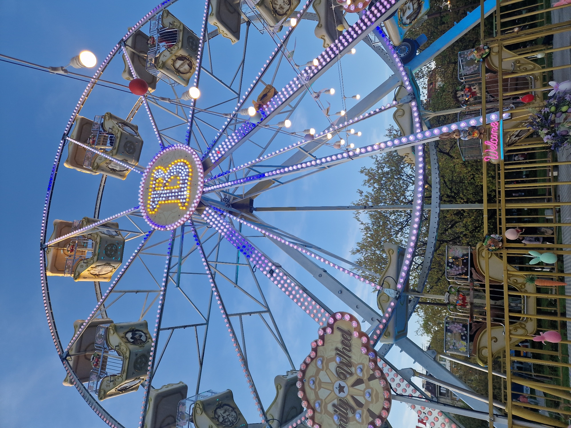 The image displays a brightly lit Ferris wheel against a clear blue sky, suggesting an amusement park or carnival setting. The large wheel is adorned with numerous purple and white LED lights along its spokes and outer rim, as well as larger, circular light bulbs. Many ornate, vintage-style gondolas or cabins are attached to the wheel, appearing ready for passengers, though no people are clearly visible within them. A prominent, stylized yellow letter "A" formed by lights is visible at the central hub of the Ferris wheel. Below this, another decorative element resembling a ship's wheel displays the text "Family Wheel."
To the right of the Ferris wheel, a golden metal fence or barrier is visible, decorated with various festive ornaments including plush bunnies, green and orange carrots, and floral arrangements, hinting at a spring or Easter theme. A pink neon sign spelling "Welcome" is also attached to this fence. A red spherical object hangs near one of the upper cabins of the Ferris wheel. Lush green trees are visible in the background, peeking through the metal framework of the attraction. The scene conveys a cheerful and inviting atmosphere, possibly during twilight or evening, given the prominence of the lights, though the clear sky suggests it could also be daytime.