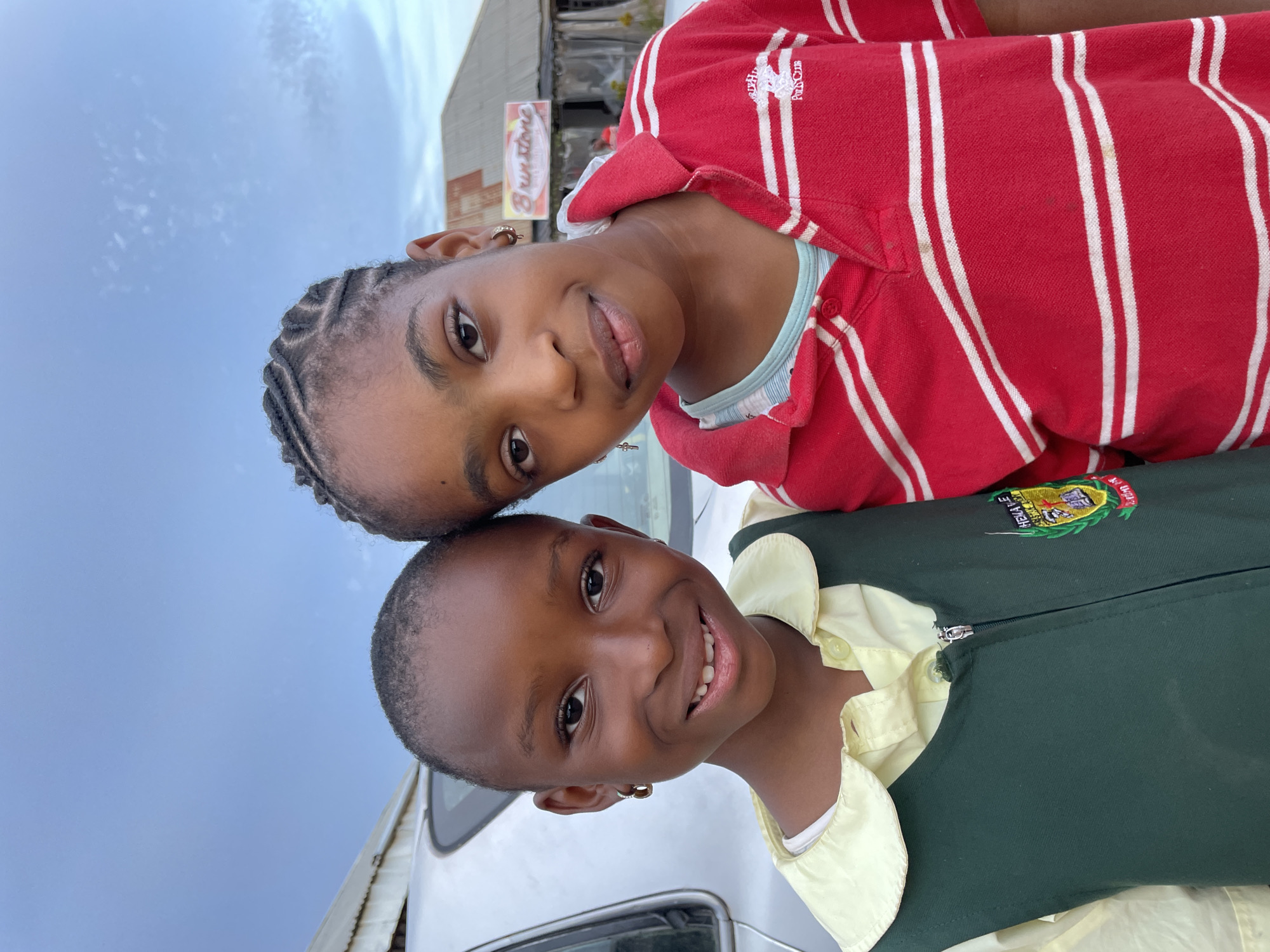 Two young, dark-skinned girls are posing closely together. The girl on top has her head tilted and her hair is styled in braids. She wears a red and white striped collared shirt. The other girl below her has a short haircut and is smiling broadly, showing her teeth. She wears a light yellow collared shirt and a dark green vest. There is a badge on the vest that says "Chenala Le."
Behind the girls is a blurred background that includes a vehicle. Above the vehicle, part of a building and the sky are visible. This is taken in Barakin Akawo, Nigeria.
