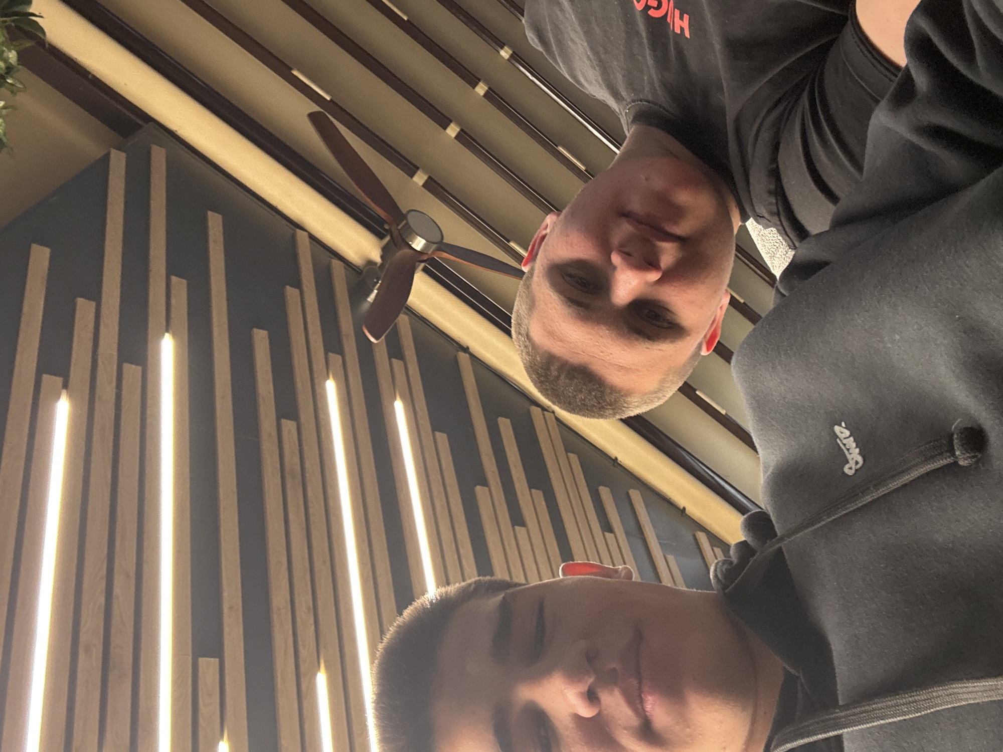 Two men are visible in a vertically oriented photograph. The background features a wall decorated with vertical wooden slats interspersed with linear light fixtures, and a ceiling with linear elements and a ceiling fan. No specific location in Targovishte, Bulgaria can be identified from the image.