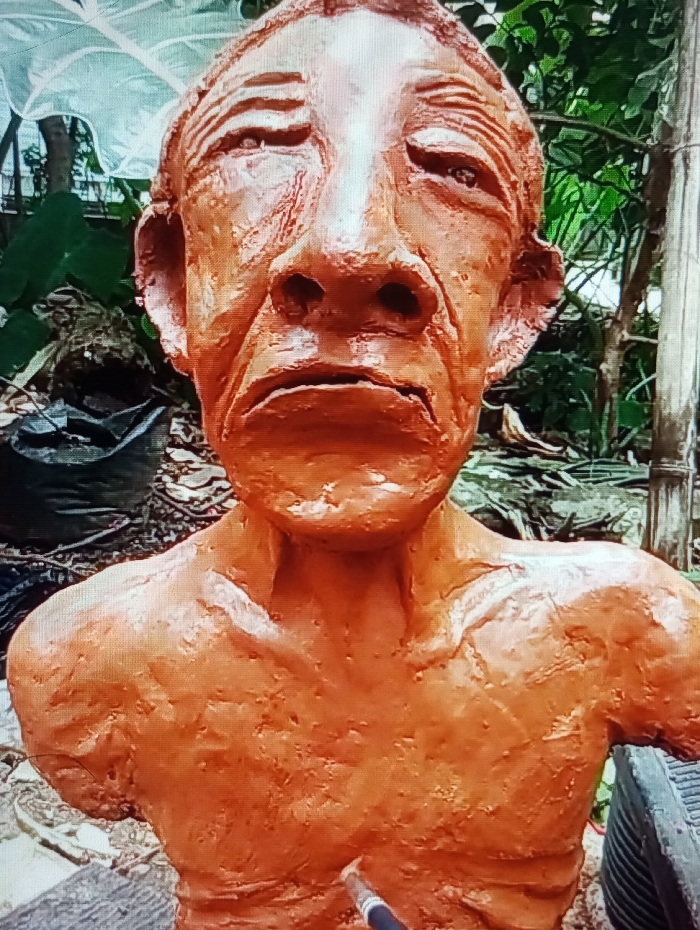 An unfinished reddish-brown bust sculpture of a human figure, crafted from a clay-like material, is depicted in an outdoor setting in Legnica, Poland. The sculpture encompasses the head and upper torso, portraying an individual with a deeply furrowed brow, closed or heavily lidded eyes, a prominent nose, and a downturned mouth, conveying a somber expression. The surface of the bust exhibits a textured, uneven quality, marked with sculpting lines. A fine-tipped brush or sculpting tool, featuring a silver ferrule and a dark handle, is positioned in contact with the lower chest area of the bust, suggesting ongoing artistic work. The background is out of focus, revealing green foliage, including a large pale green leaf in the upper left, dark ground cover, and a vertical pole on the right side.