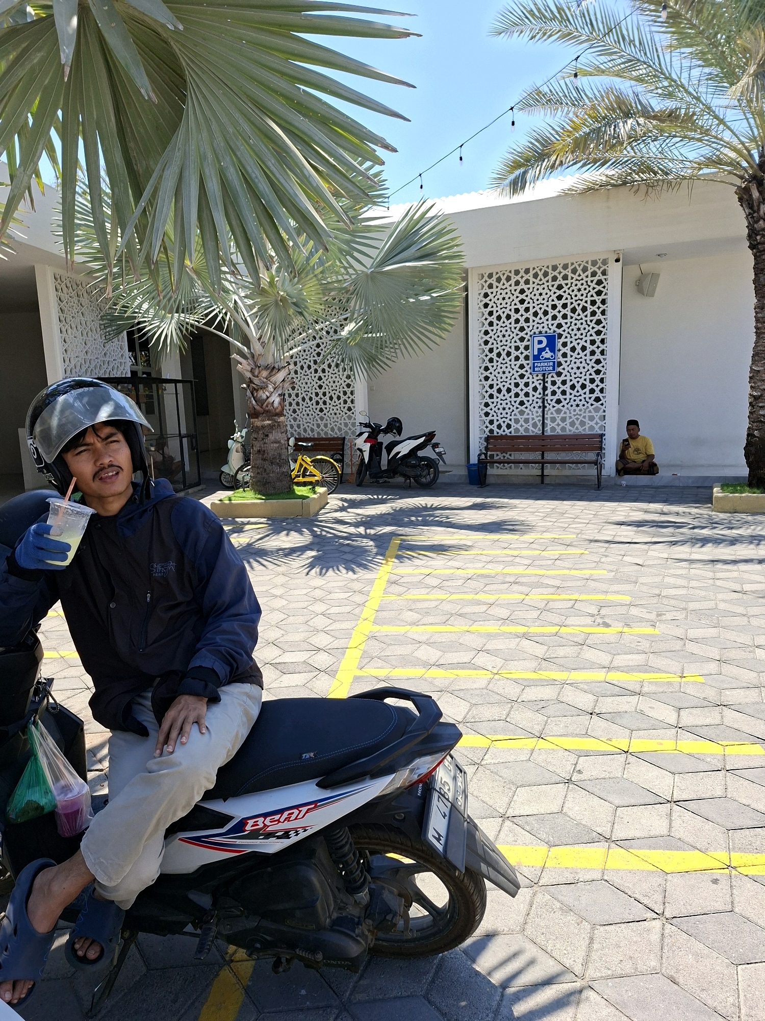 The image depicts an outdoor scene on a sunny day, likely in a parking area, with elements suggesting a location in Branta Tinggi, Indonesia. In the foreground, a young man wearing a helmet and gloves is seated on a white and black motorcycle. He holds a clear plastic cup with a straw, containing a light-colored beverage. He is dressed in a dark jacket and light-colored pants. The motorcycle has a green plastic bag hanging from its side.
The background features a white building with intricate white lattice work on its facade. Adjacent to the building is a wooden bench. A blue sign with a white "P" and a motorcycle symbol, labeled "PARKIR MOTOR," is visible. To the right of the bench, a man in a yellow shirt and sarong is seated on the ground, looking at his mobile phone.
The ground is paved with hexagonal tiles, marked with yellow lines indicating parking spaces. Several motorcycles are parked in the area, including one yellow bicycle. Palm trees, with their fronds casting shadows on the ground, are prominent in the scene, suggesting a tropical or subtropical climate. The sky is clear and blue. The overall atmosphere appears relaxed and casual.