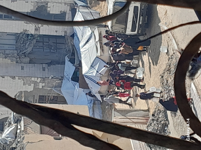 The image depicts a crowded, damaged urban environment, specifically identified as the city of Khan Yunis, Palestinian Territory. The scene is observed through a narrow, irregular frame, possibly from an interior or through debris.
In the background, multiple multi-story buildings are severely damaged, displaying large structural breaches, exposed interiors, and peeling facade material, suggesting significant destruction. Below these damaged structures, numerous makeshift shelters fashioned from white tarpaulins, some with blue trim, are erected on sandy, unpaved ground.
The foreground and midground are populated by a large group of individuals, appearing to be mostly adults and children, dressed in a mix of casual and traditional attire. A central activity involves a white Volvo truck, around which a crowd has gathered. Many individuals are carrying or are in possession of clear plastic jerry cans or water containers. People are seen actively engaged in collecting or transporting these containers, indicative of water distribution. One person, a young individual in a dark shirt and shorts, carries two such containers. Another individual in a black abaya sits on a pile of rubble in the lower-left. Piles of construction debris and rubble are scattered across the sandy terrain. The overall environment appears to be one of displacement and resource scarcity. The lighting indicates daylight.