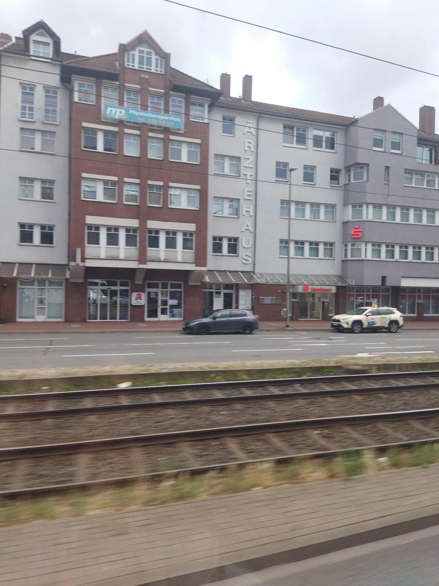 The image depicts a street scene in Hanover, Germany, with a multi-story building dominating the background. The building features a mix of brick and grey facades, with numerous windows. Signs are visible on the building, including one in blue and white with "mp" and "Physiotherapie Puls" text, another with vertical lettering that reads "ÄRZTEHAUS", and a red logo that appears to be for Sparkasse, a German savings bank.
In the foreground, a street runs horizontally across the image, with two cars visible. A dark-colored sedan is parked on the left side of the street. On the right side, a white SUV, possibly a taxi, is driving. In front of the street, train tracks are visible, with ballast and vegetation alongside them. The sky is overcast, suggesting a cloudy day. The overall impression is of an urban environment with commercial and residential buildings lining a street with an adjacent railway line.