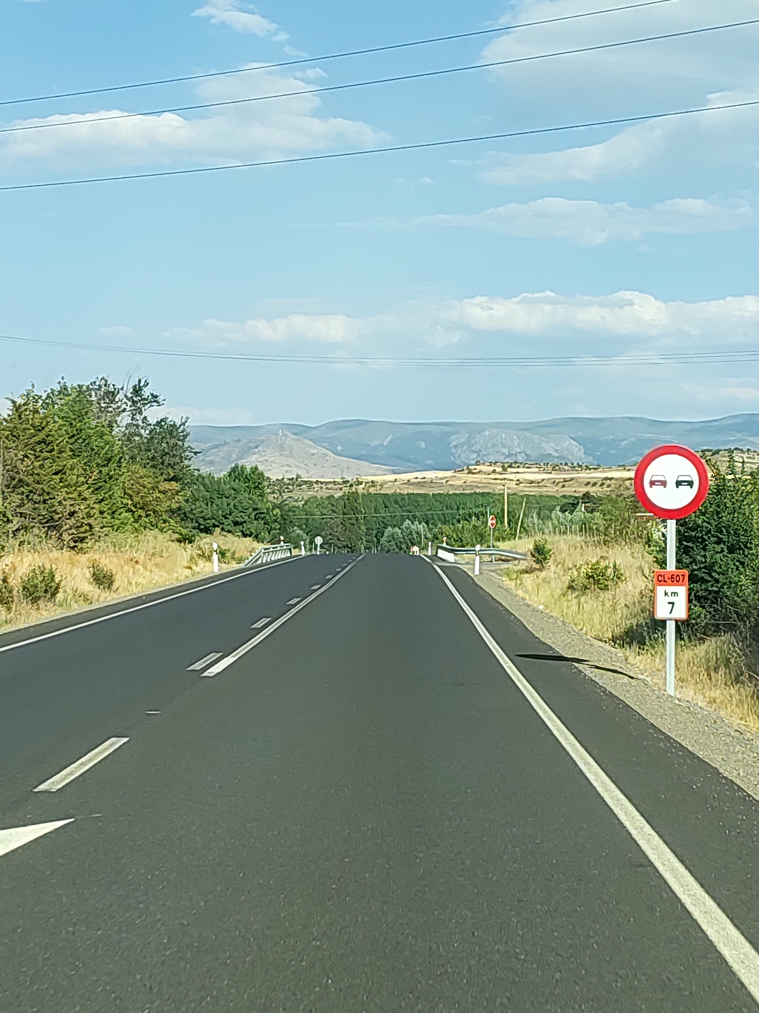 The image depicts a two-lane asphalt road stretching into the distance under a bright, cloudy sky. The road has white lane markings. To the right of the road, there is a sign with a red circle and a diagonal bar, indicating a prohibition. Inside the circle are two car symbols, one in front of the other, suggesting a restriction on overtaking. Below this sign is a smaller rectangular sign with "CL-607", "km", and the number "7" displayed, indicating a road marker. The surrounding landscape is dry and rural, with sparse vegetation, including bushes and trees. In the background, rolling hills and mountains are visible. The time of day appears to be daytime, likely late morning or early afternoon, given the strong sunlight and shadows. There are no people or vehicles clearly visible in the scene, besides the implied presence of the overtaking restriction. The scene is characteristic of a rural highway in the province of Segovia, Spain, given the "CL-607" designation, which is a regional road in Castilla y León.