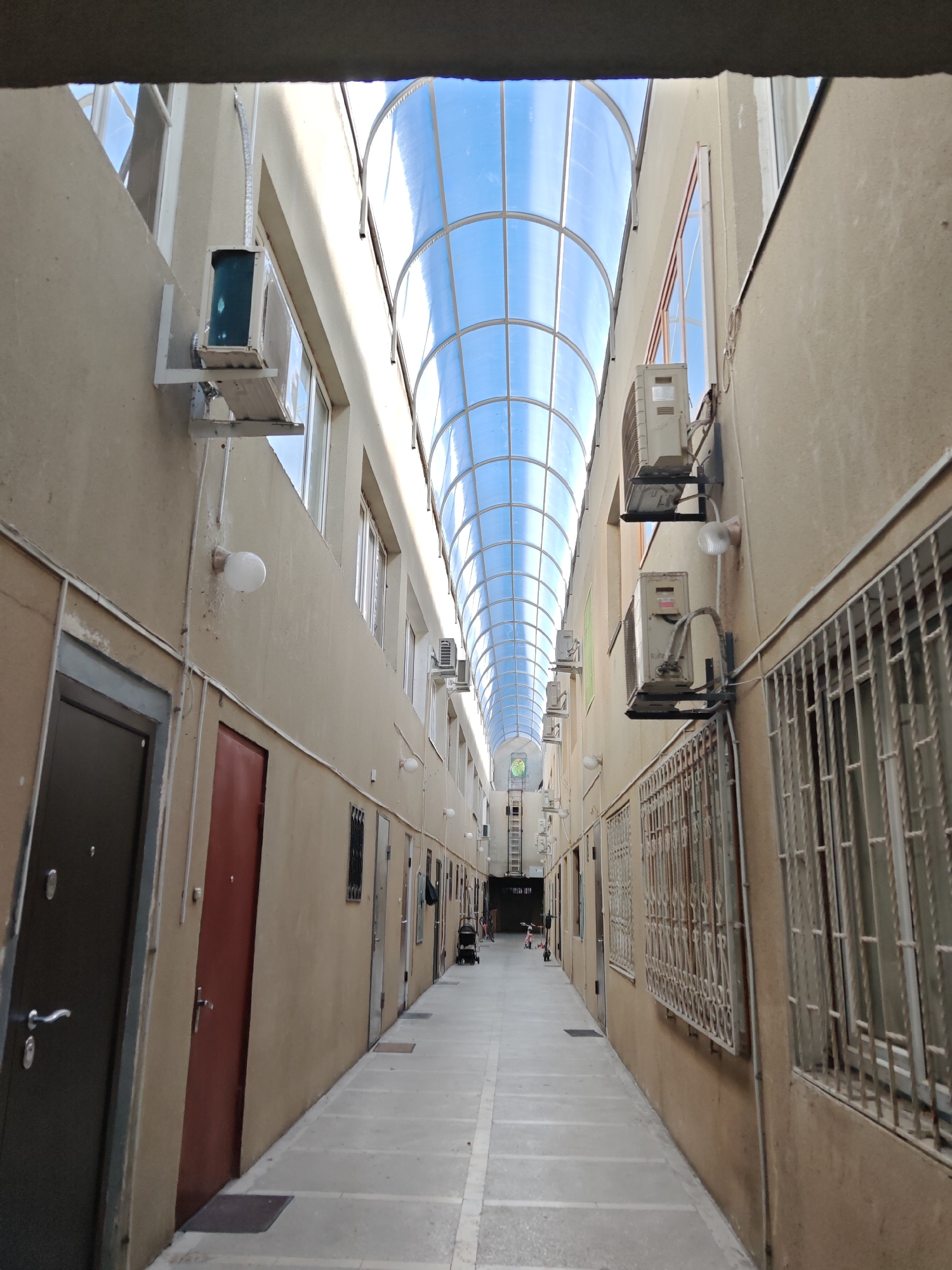 The image captures a long, narrow outdoor corridor or passageway within a building complex in Sochi, Russia. The passageway is covered by a translucent, arched roof made of clear panels, through which a bright blue sky is visible, indicating daytime.
On both sides, the walls of the buildings are a light tan or beige color. Numerous entry doors, varying in shades from dark brown to reddish-brown, line the walls, suggesting individual units or apartments. Many windows are also visible, some fitted with white metal security grilles.
A prominent feature is the array of outdoor air conditioning units mounted at different heights on both walls, highlighting the need for climate control in the environment. Spherical white light fixtures are spaced along the walls to provide illumination. The ground is a paved walkway, leading into the distance.
Towards the far end of the corridor, the perspective narrows, revealing more doors, windows, and air conditioners. In the distance, a child's scooter and a stroller are faintly discernible on the walkway, hinting at the presence of residents or activity, though no clear individuals are depicted. A ladder is visible against a far wall.
The scene portrays a functional, residential or mixed-use walkway, brightly lit by natural light filtering through the overhead structure. The overall impression is one of an urban pathway adapted for comfort and security in a warm climate. No visible text is clearly legible in the image.