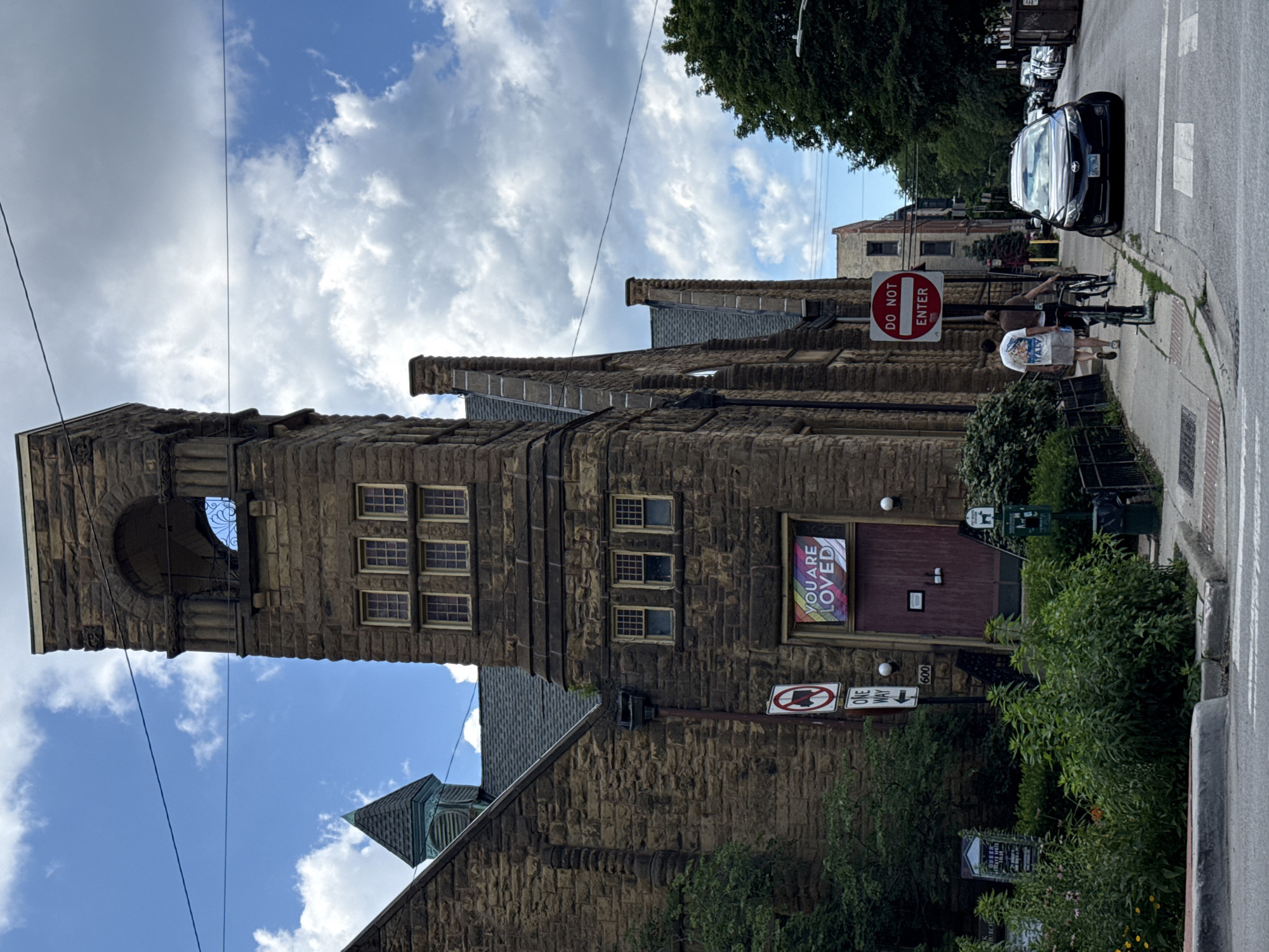 A stone building, possibly a church, is the primary subject of this image, which is oriented vertically. The building features a tall, arched tower on the left and multiple windows on the upper levels. A banner with the words "YOU ARE LOVED" in rainbow colors is displayed in one of the windows. Several signs are visible, including a "DO NOT ENTER" sign with a red circle and slash, and a "ONE WAY" sign. A black car is parked on the right side of the street. Two people are walking on the sidewalk near the car and the "DO NOT ENTER" sign. The sky above is partly cloudy, suggesting it is daytime. Lush green bushes and trees surround the building. The location is identified as Chicago, USA.