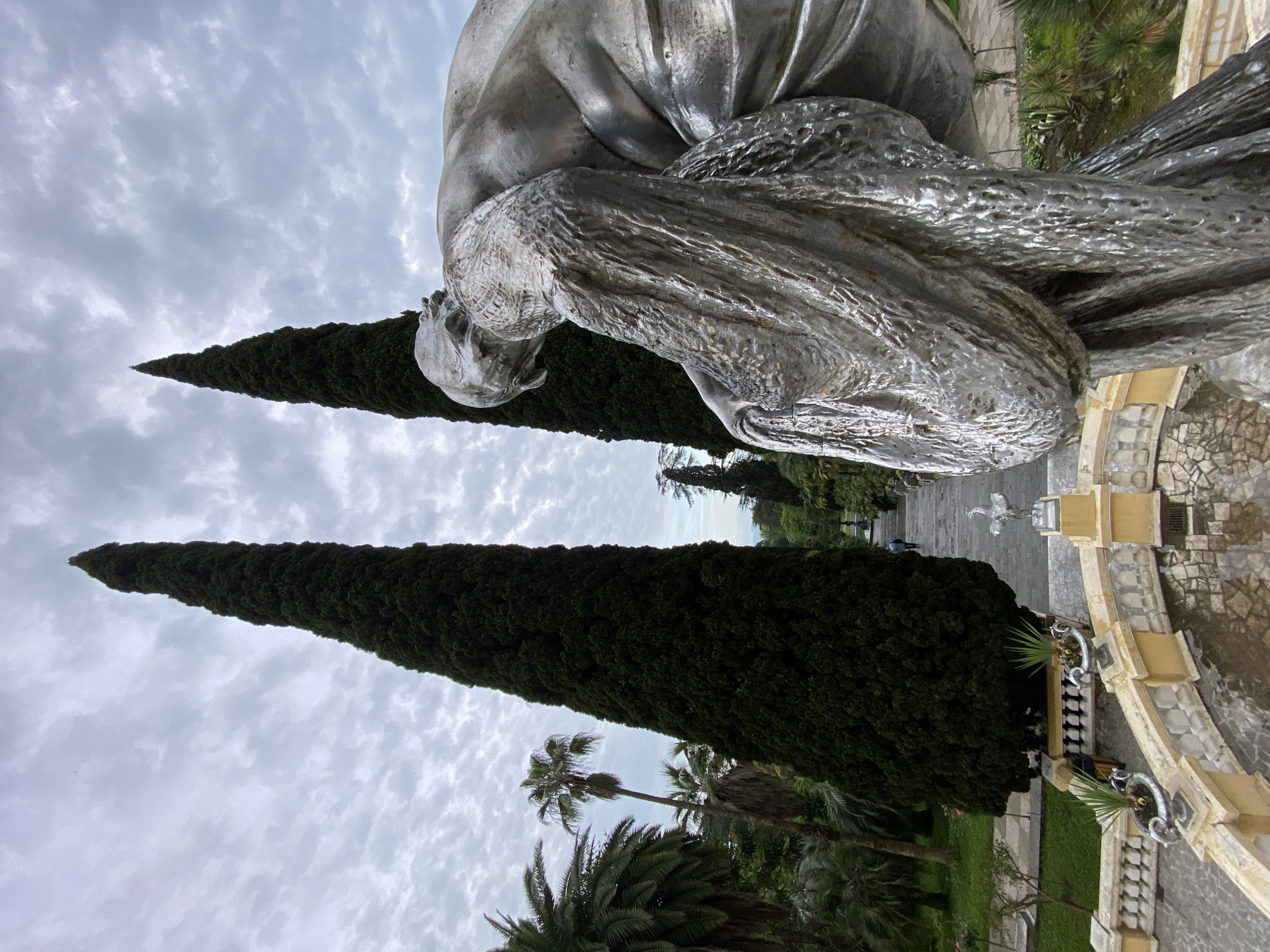 This image shows a statue in a park in Sochi, Russia. The statue is of a human figure, sculpted in a reflective metal material, potentially depicting a moment of sorrow or contemplation. The statue is positioned near two tall, narrow evergreen trees that draw the eye upward toward the cloudy sky. In the background, there's a paved walkway leading to a fountain, adding depth to the scene. The edge of an ornate, yellow-toned stone structure with balustrades is visible, bordering the park's edge. The presence of palm trees indicates a subtropical climate typical of Sochi.