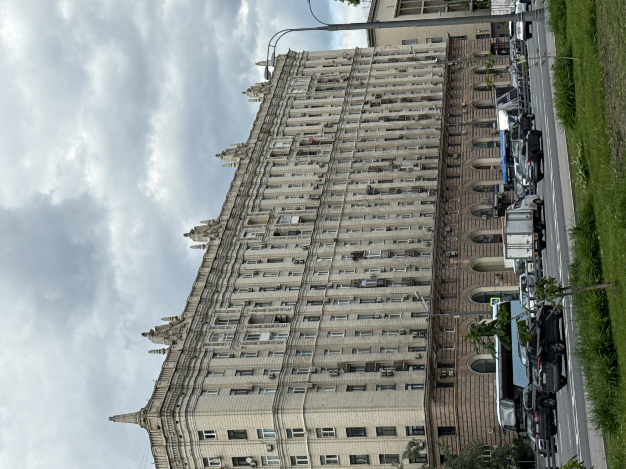 An outdoor, slightly rotated shot captures a grand, multi-story building under an overcast sky. The prominent subject is a large, ornate building, characteristic of Stalinist Empire style architecture often found in Moscow, Russia. Its facade is a light, possibly cream-colored stone, featuring numerous rectangular windows uniformly arranged across many floors. The building is heavily embellished with decorative vertical and horizontal elements, including cornices and multiple spire-like pinnacles along its roofline. Many windows have externally mounted air conditioning units. The lower portion of the building, visible at the bottom of the structure, appears to be constructed from a darker, textured stone with distinct arched windows or doorways.
Adjacent to the building is a street, lined with numerous parked vehicles, including cars and at least one truck, suggesting an urban setting. A strip of green grass borders the street, adding a touch of nature to the concrete environment. A lamppost extends into the frame from the right side, its arm curving over the street and part of the building. The sky is heavily overcast with dense, grey clouds, indicating a cloudy or gloomy day. No people are visible in the scene. There is faint, illegible text on the building's facade, possibly a name or historical marker, and a small, unreadable sign is visible near the parked cars.